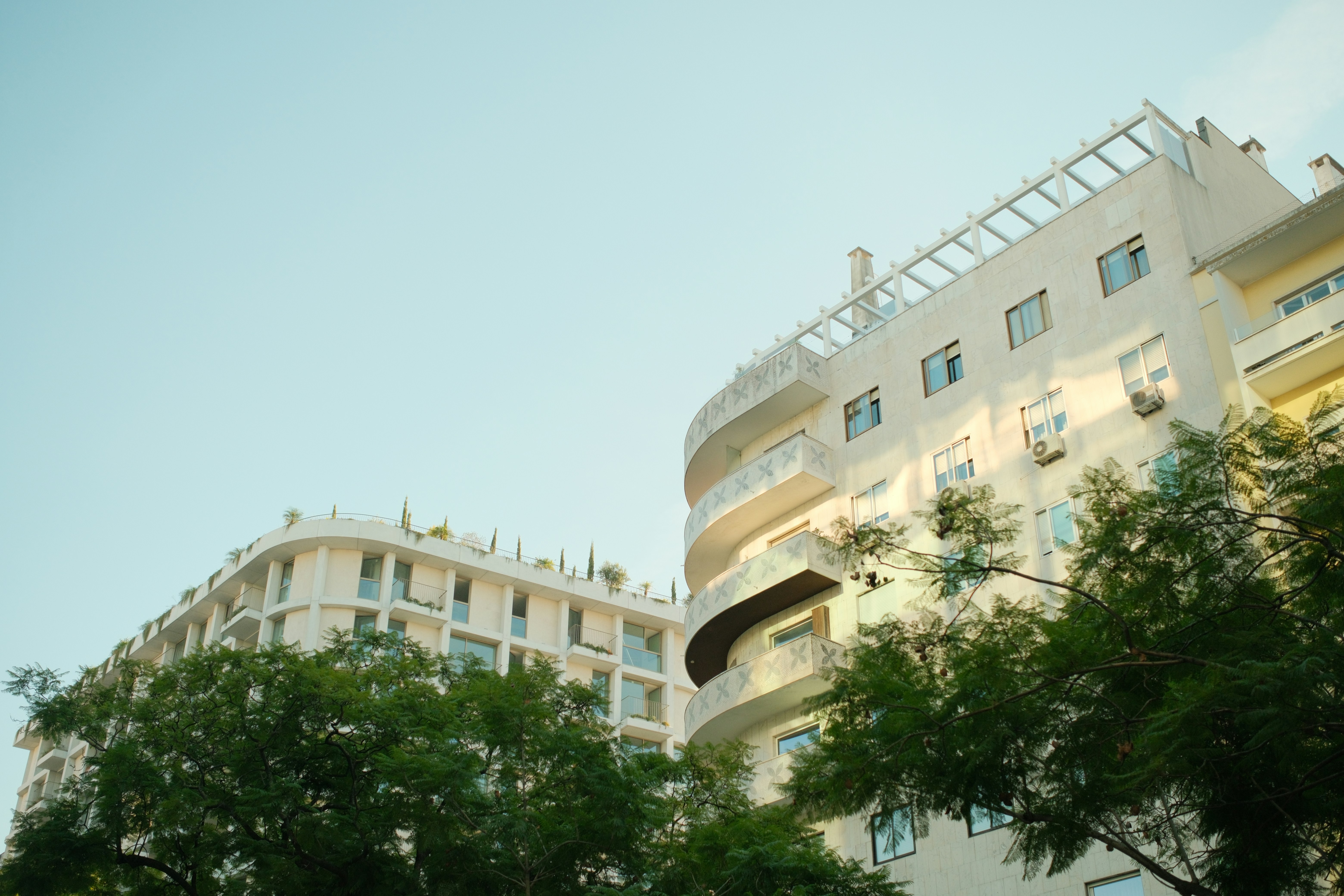 Modern white apartment buildings with curved balconies and green trees against a clear sky