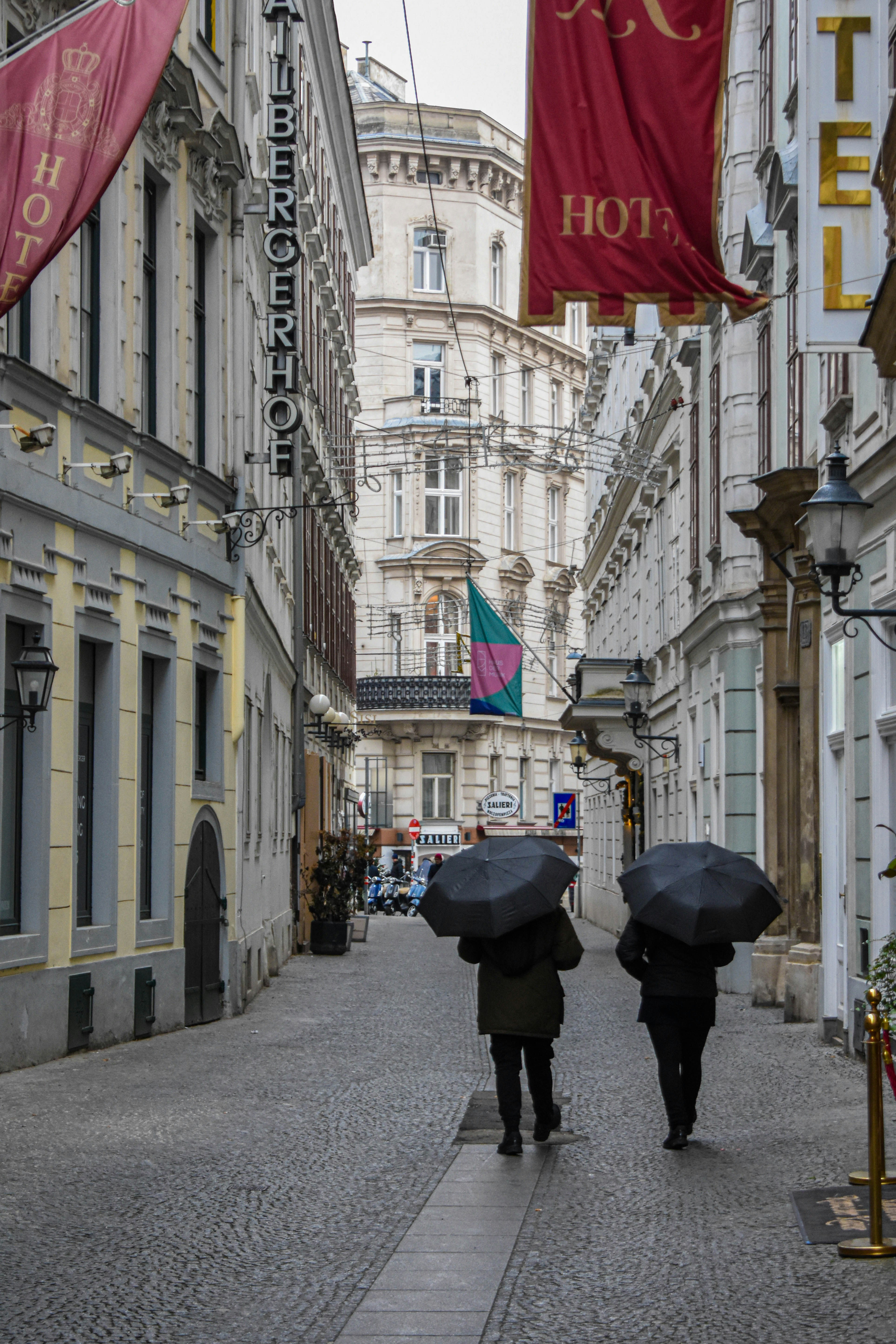 Two people with black umbrellas walking down a cobblestone street in Vienna