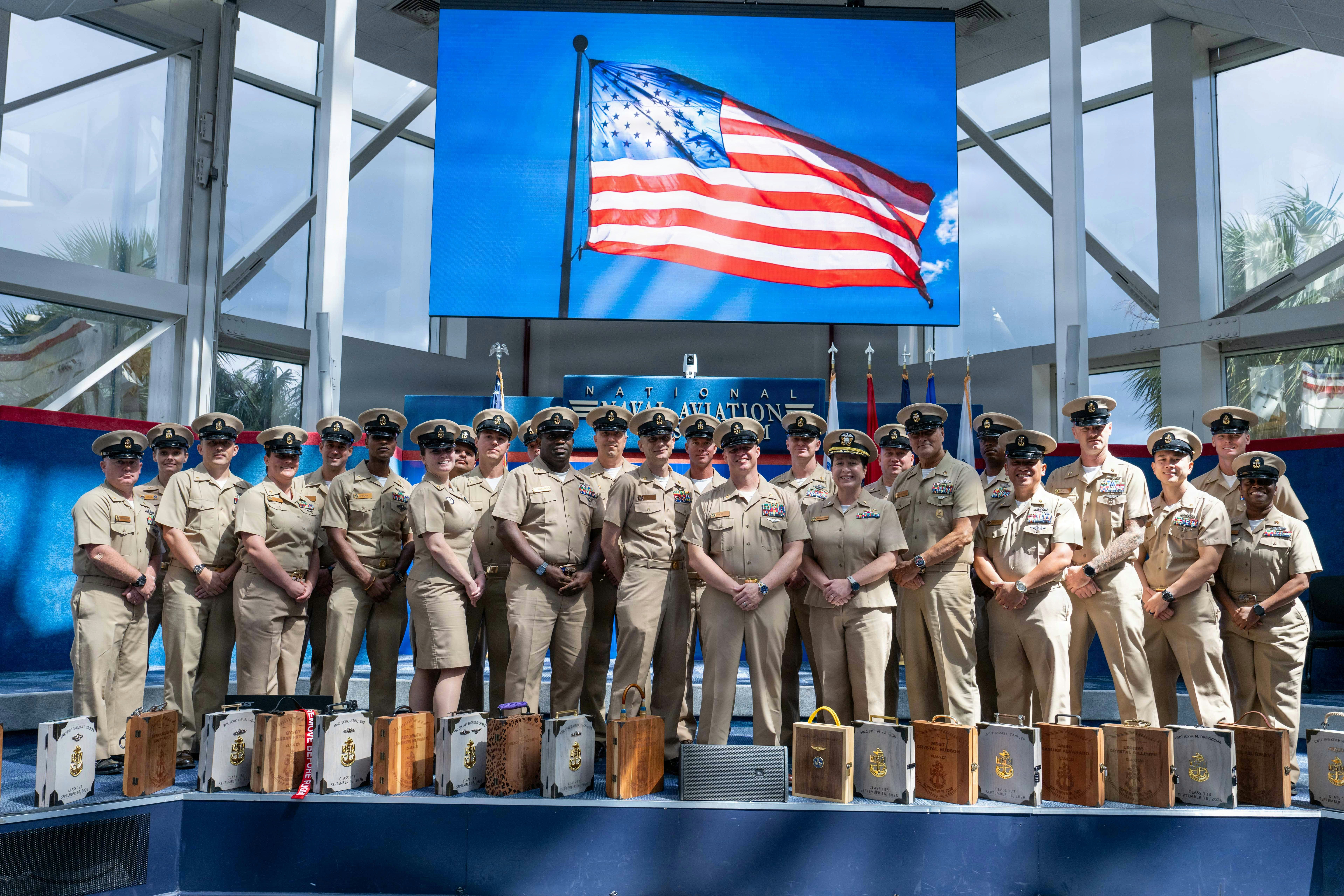 A group of US Navy personnel in khaki uniforms standing before a large American flag