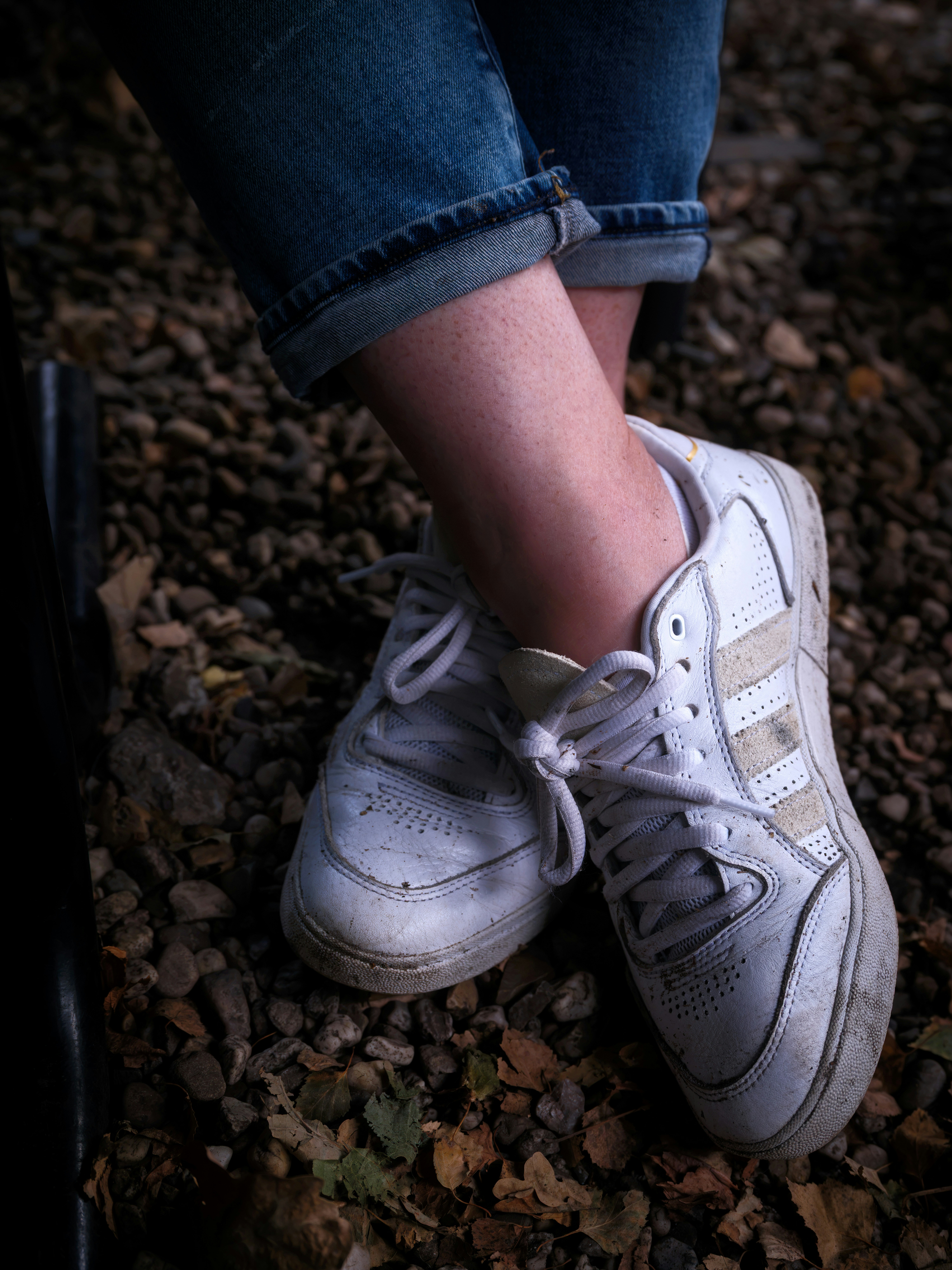 A person wearing blue jeans and dirty white sneakers on a gravel path