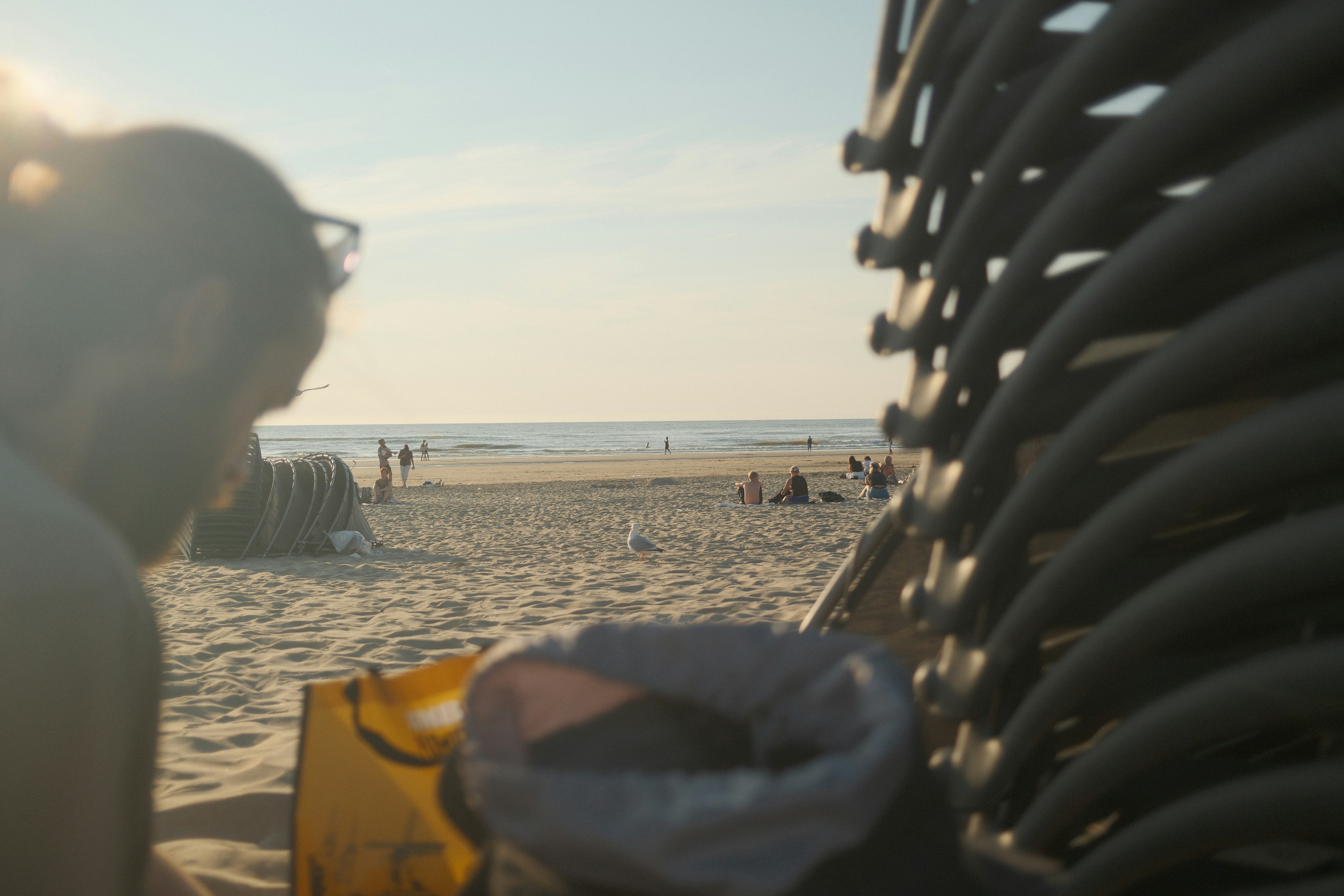 A person in the foreground overlooking a sandy beach with people and stacked chairs