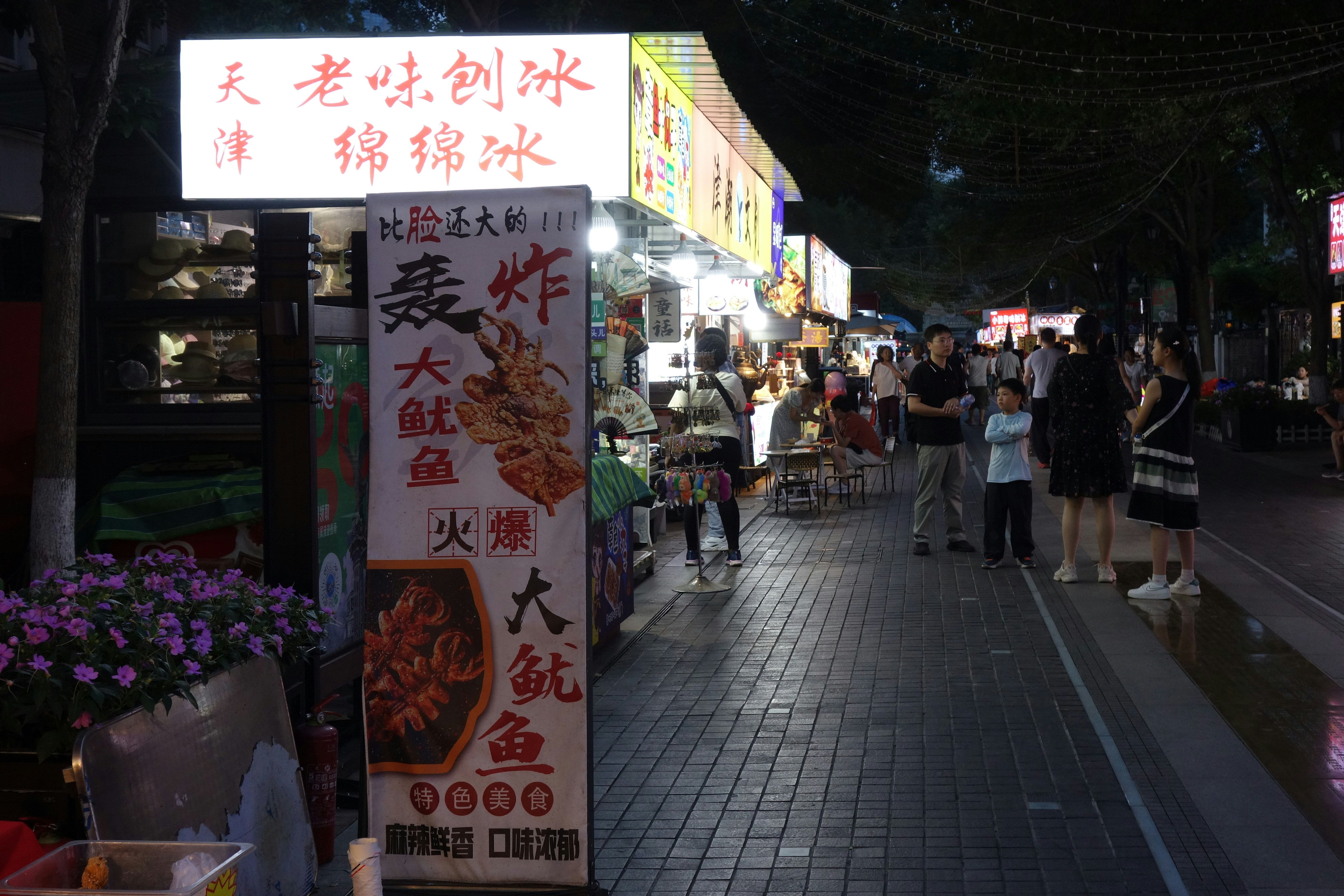 People walking along a night market street with illuminated food stalls and signage