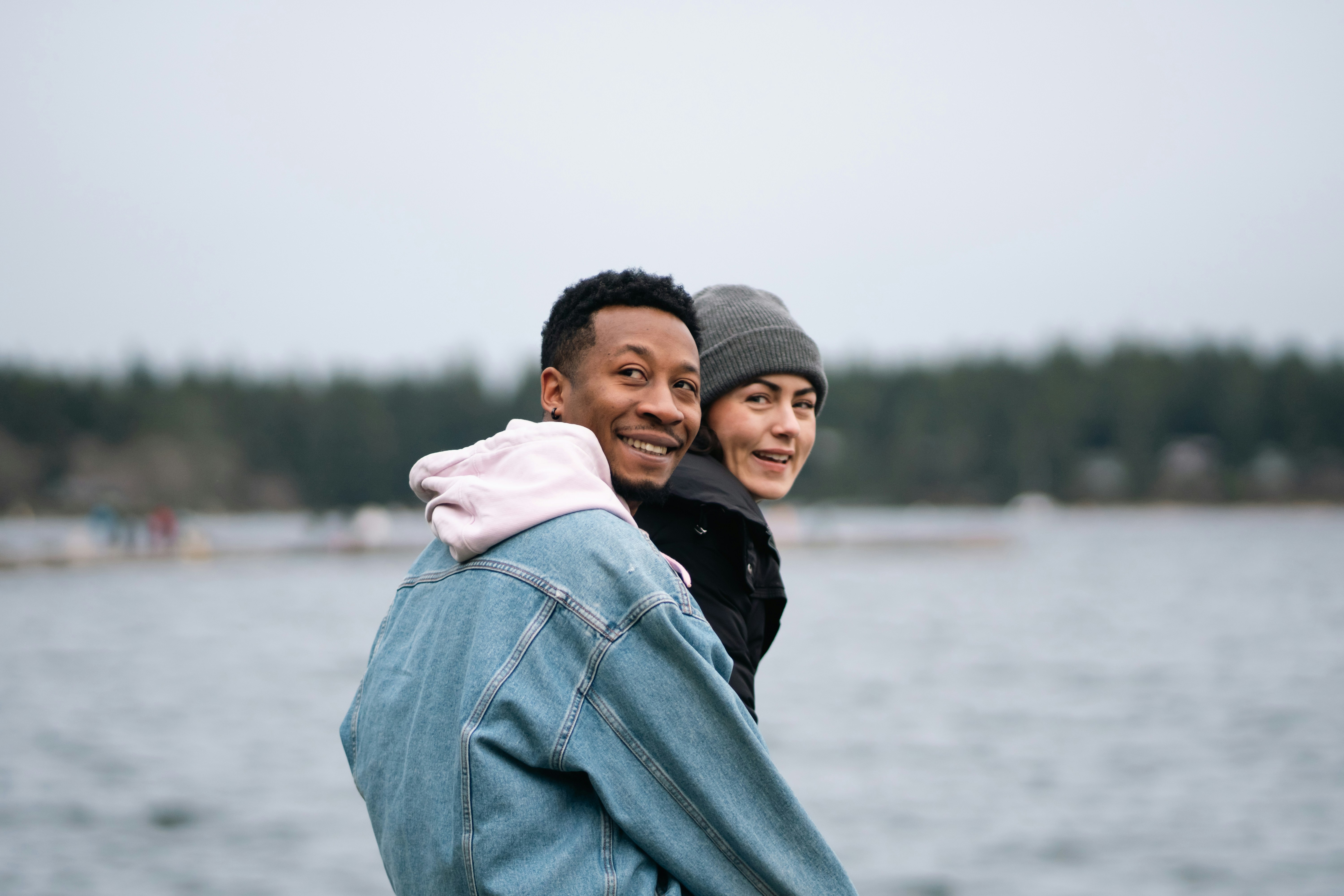 A man in a denim jacket and a woman in a beanie by a lake