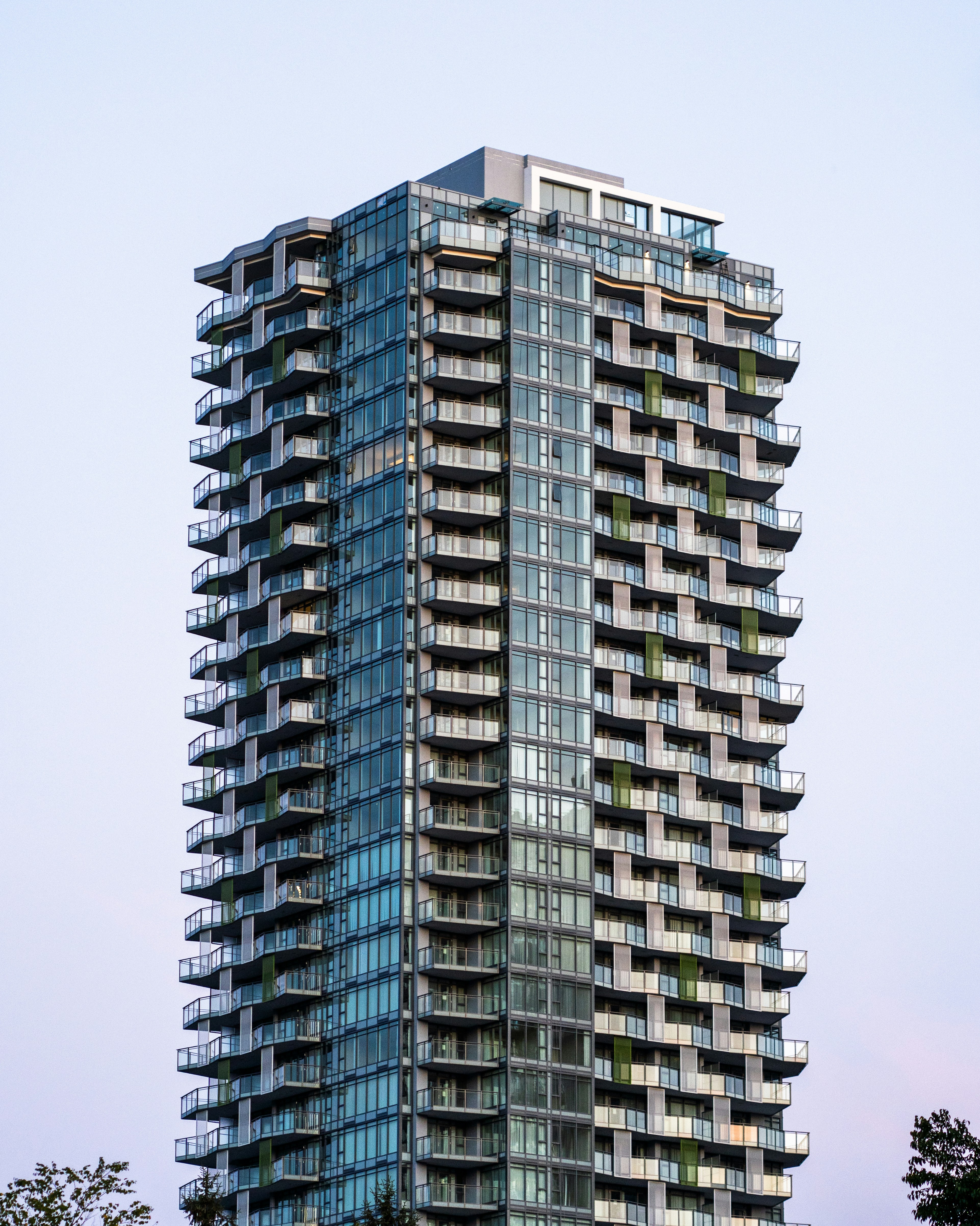 A tall modern glass skyscraper with many balconies against a pale sky