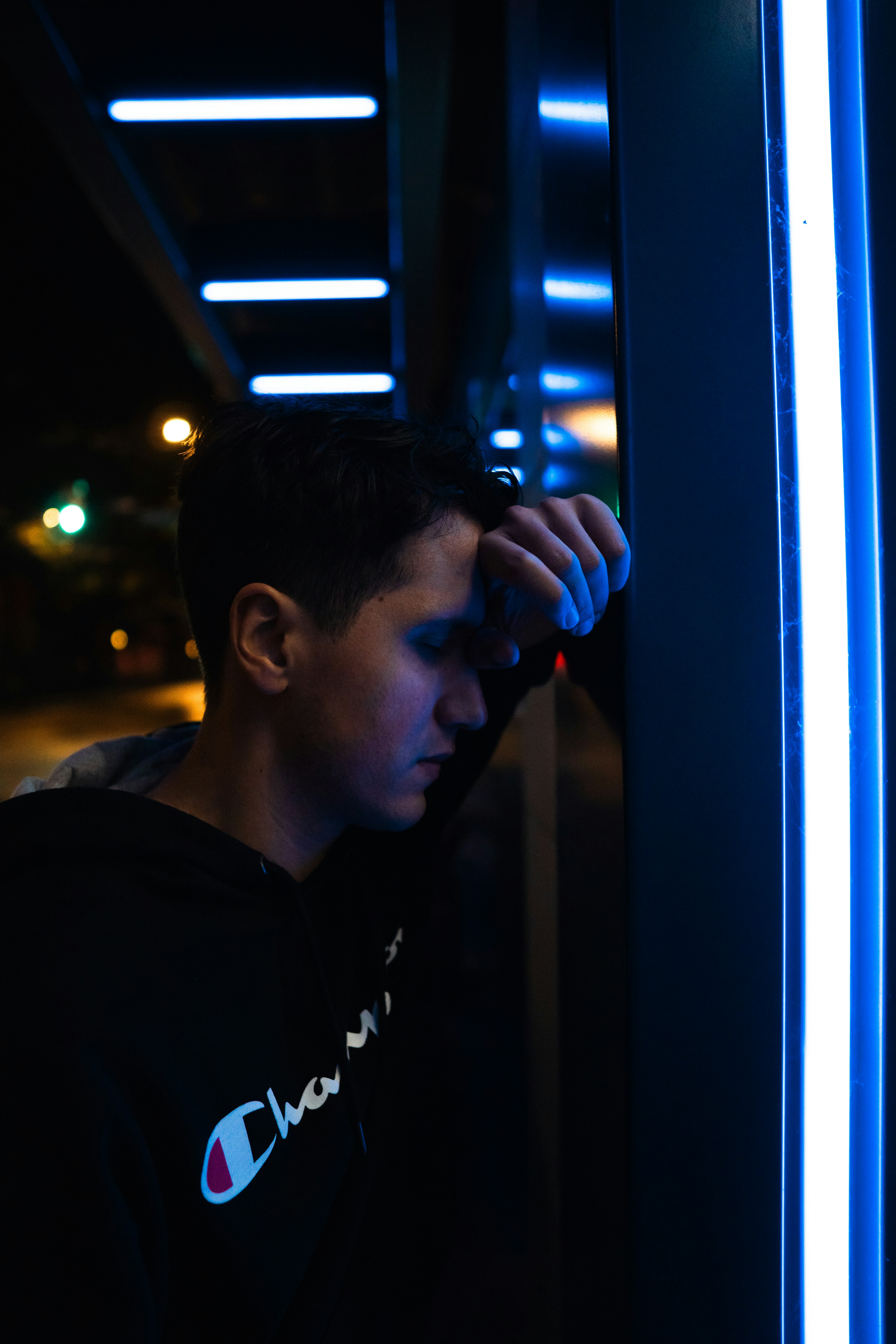 A person in a black hoodie leaning against a wall with blue neon lights
