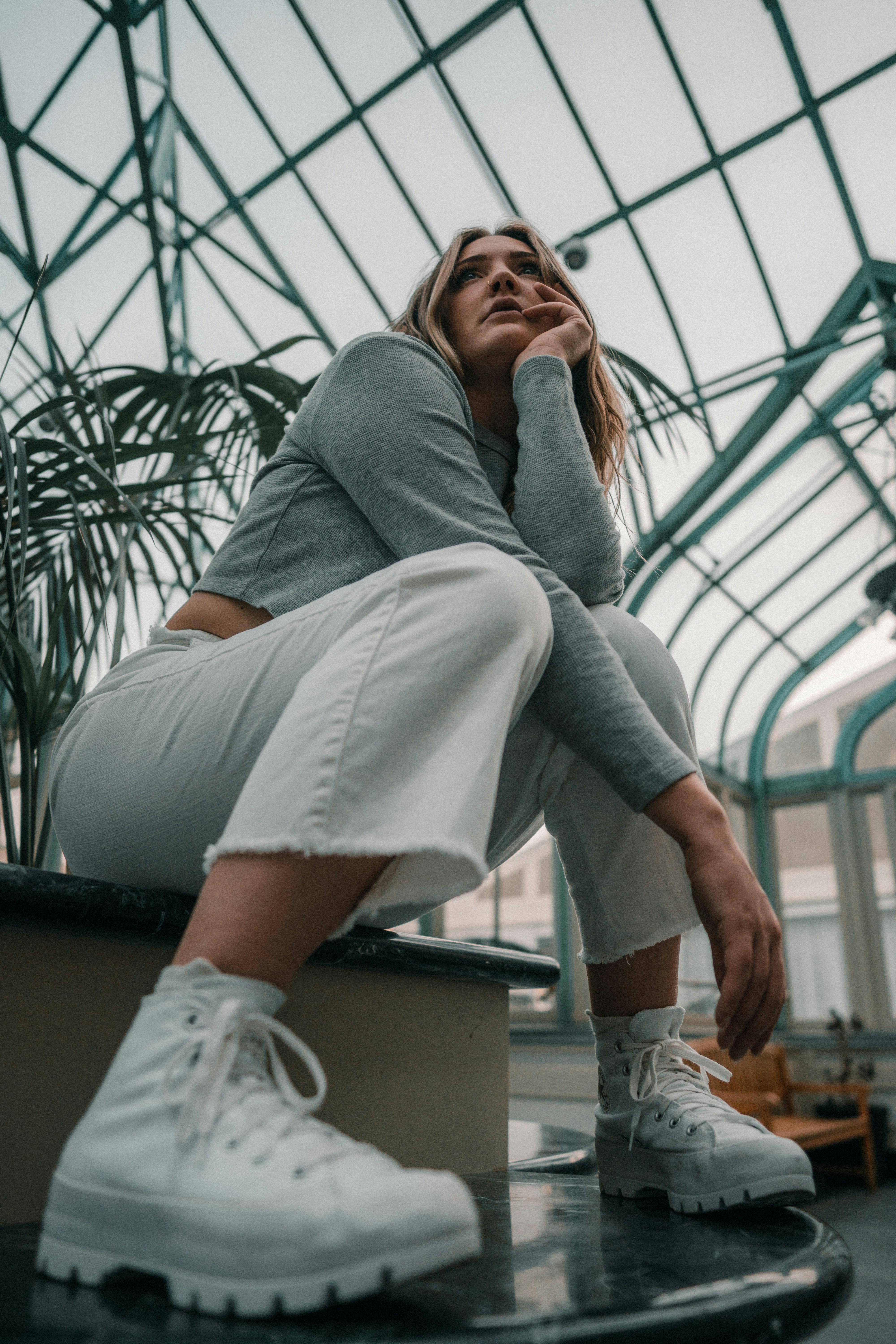 A woman in a grey long-sleeved shirt and white jeans in a glass conservatory