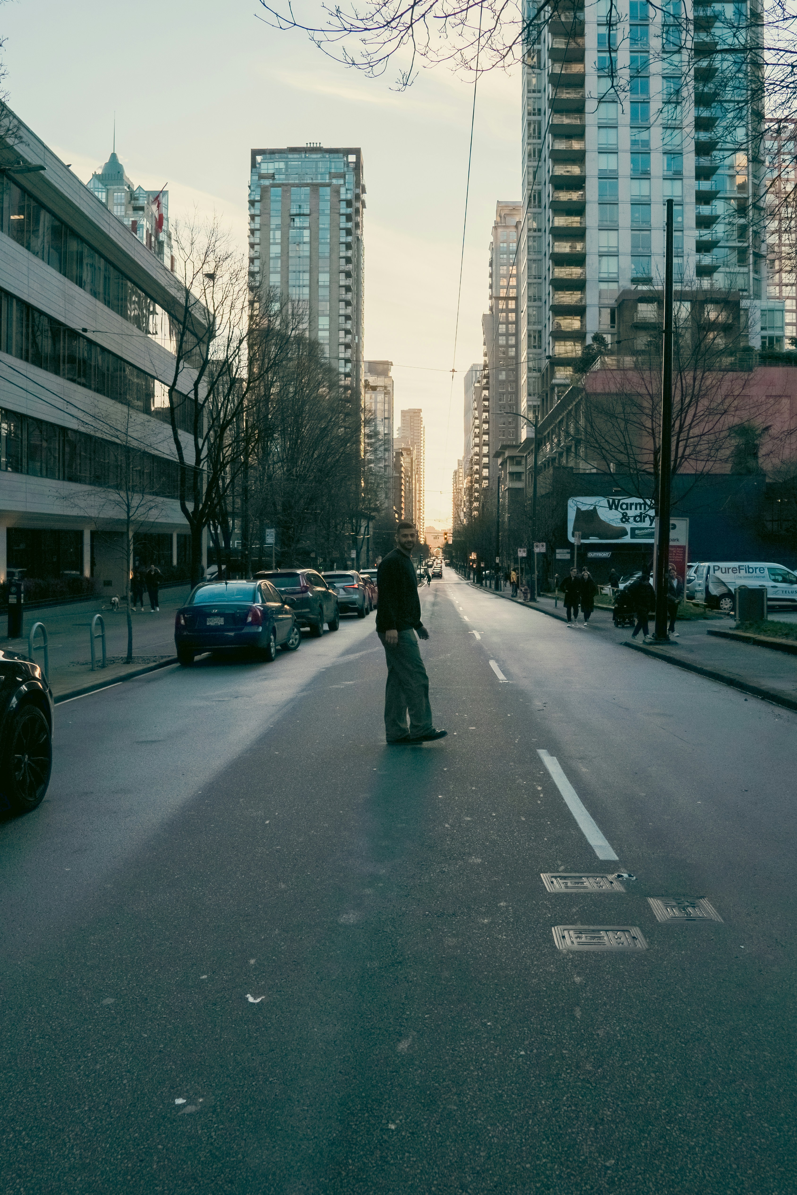 A man walking across a city street between tall buildings in low light