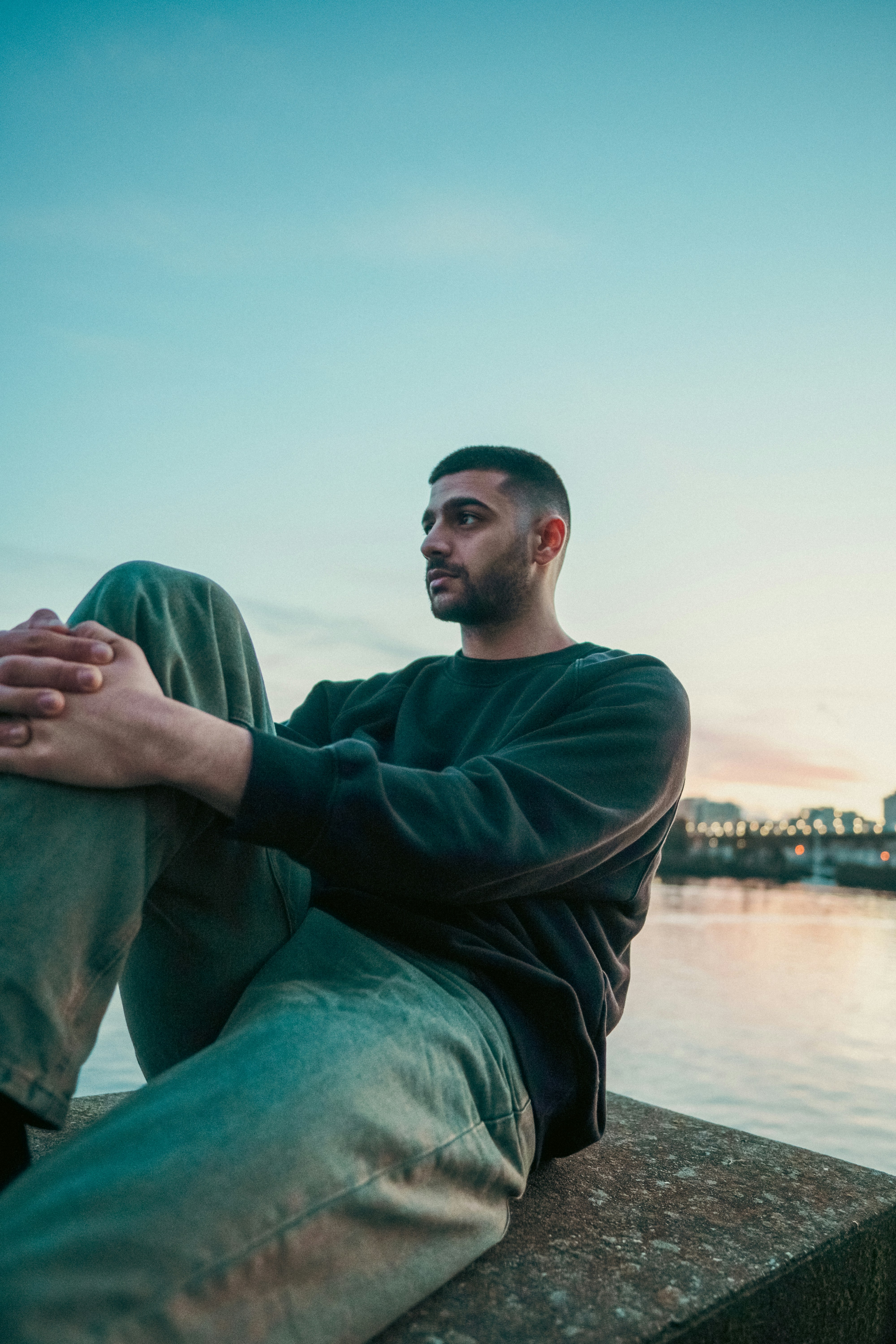 A man in a dark sweatshirt sitting on a concrete ledge by water