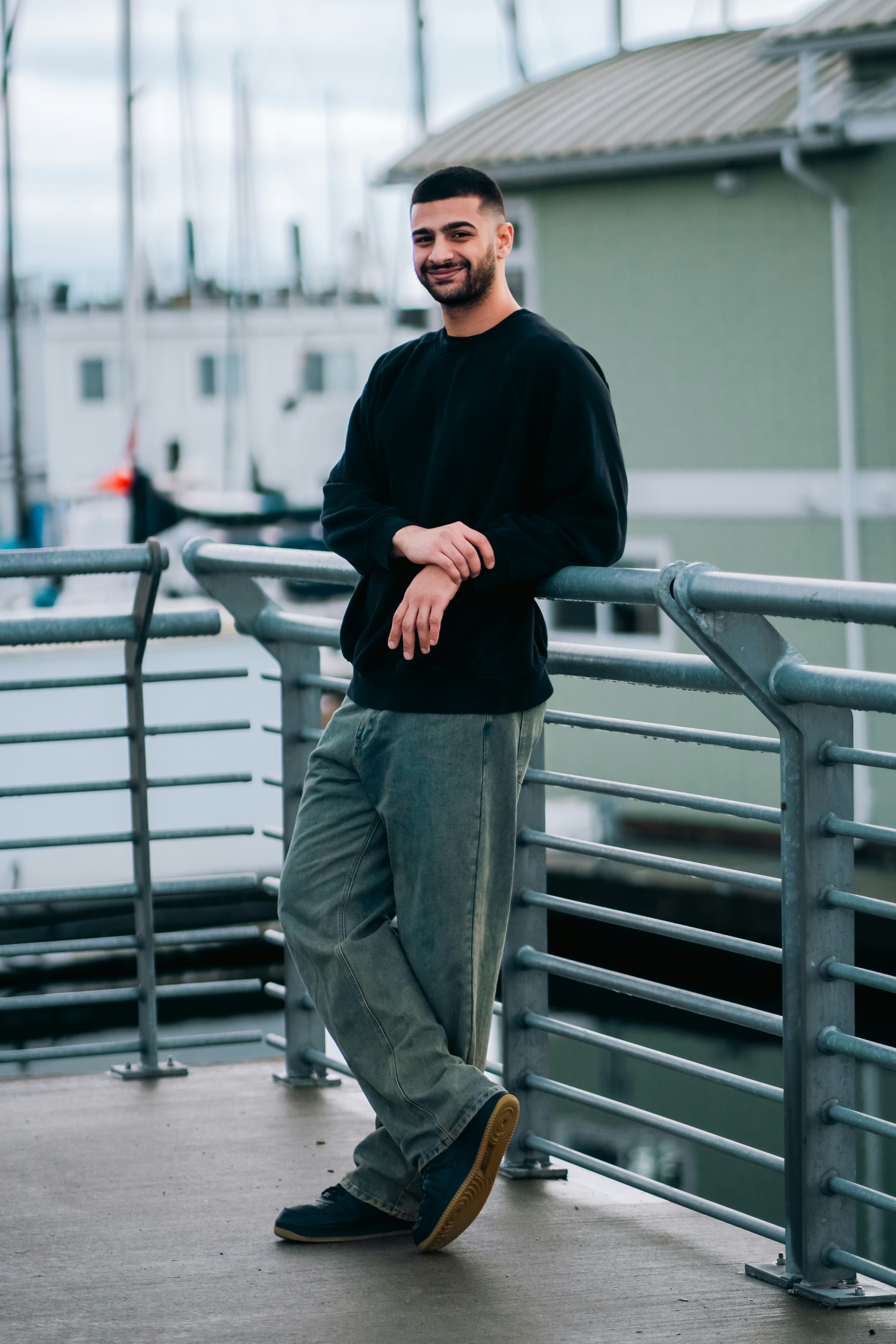 A man in a black sweatshirt and grey jeans leaning against a metal railing