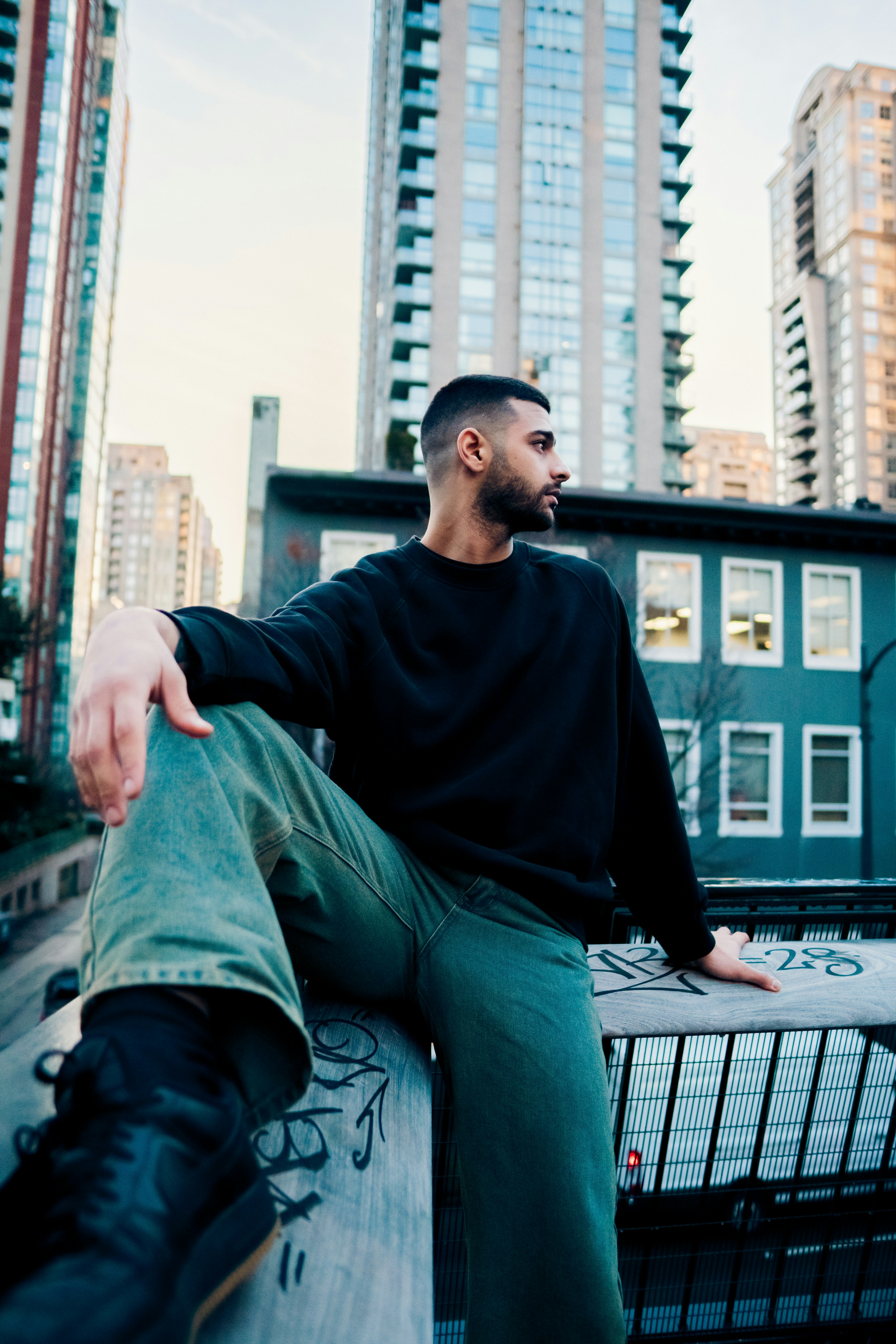 A man in a black sweatshirt and green jeans sitting on a graffiti-covered railing