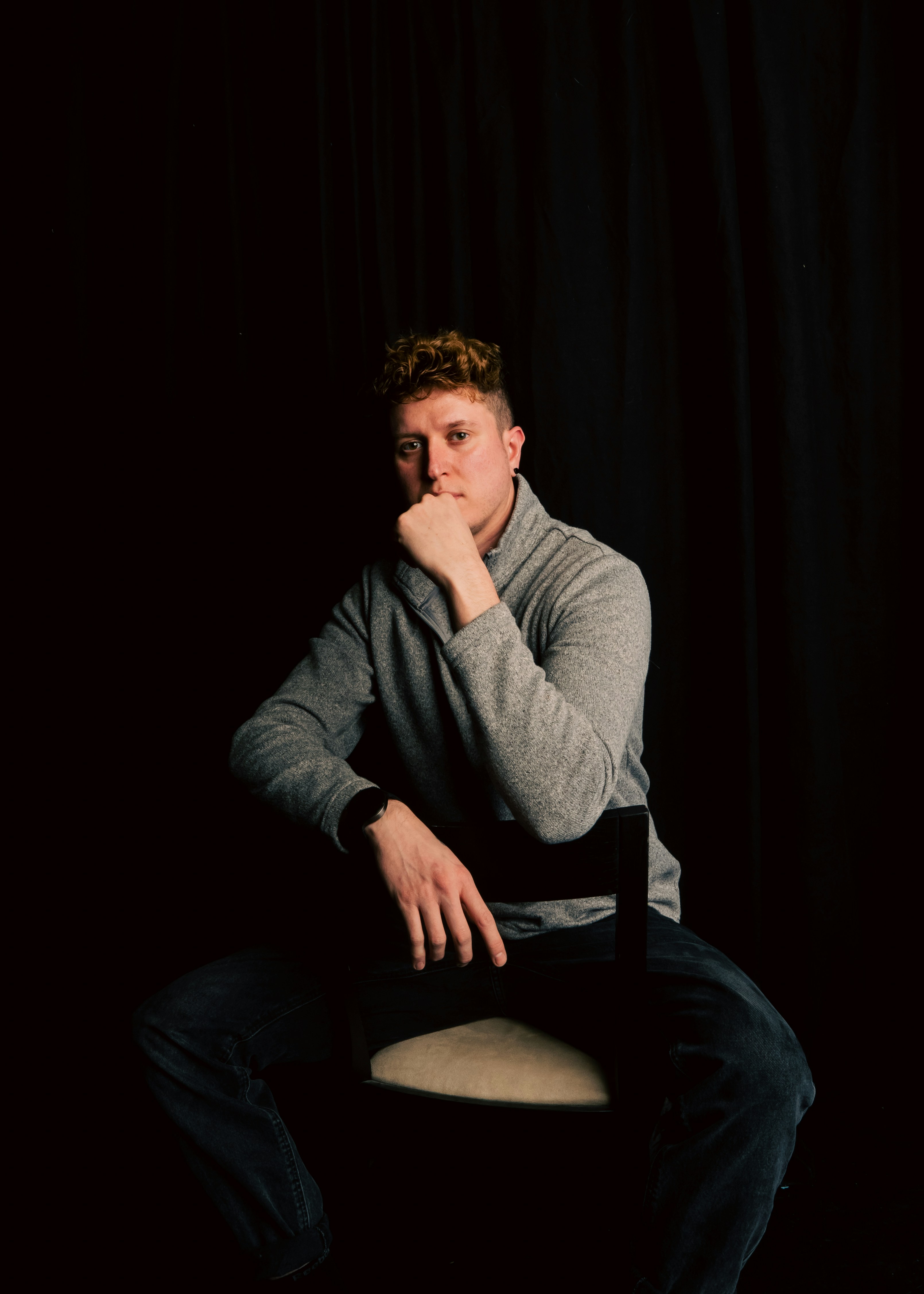 A man with curly red hair in a grey sweater sitting on a chair