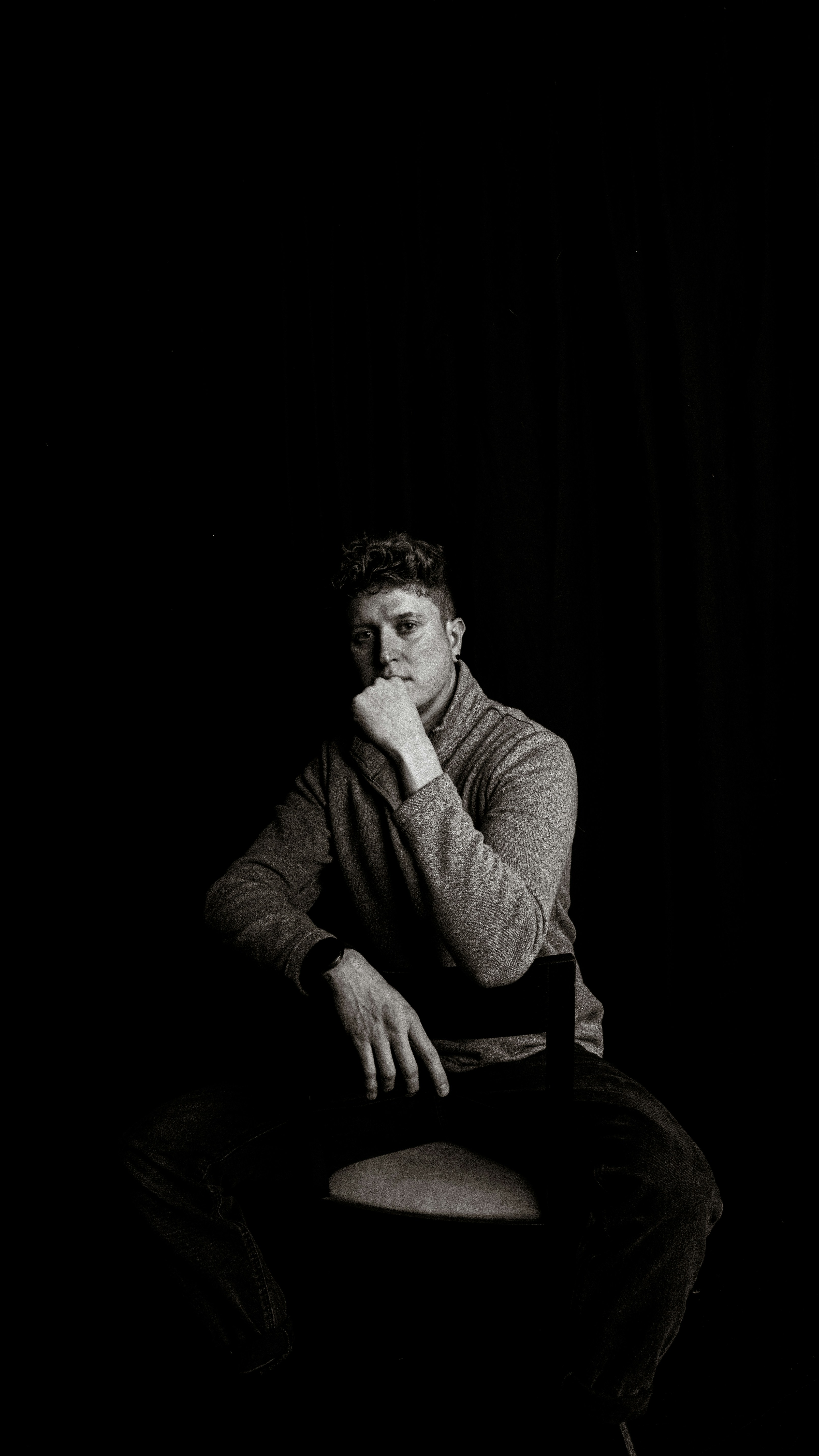 A man in a grey sweater sitting on a chair against a black background