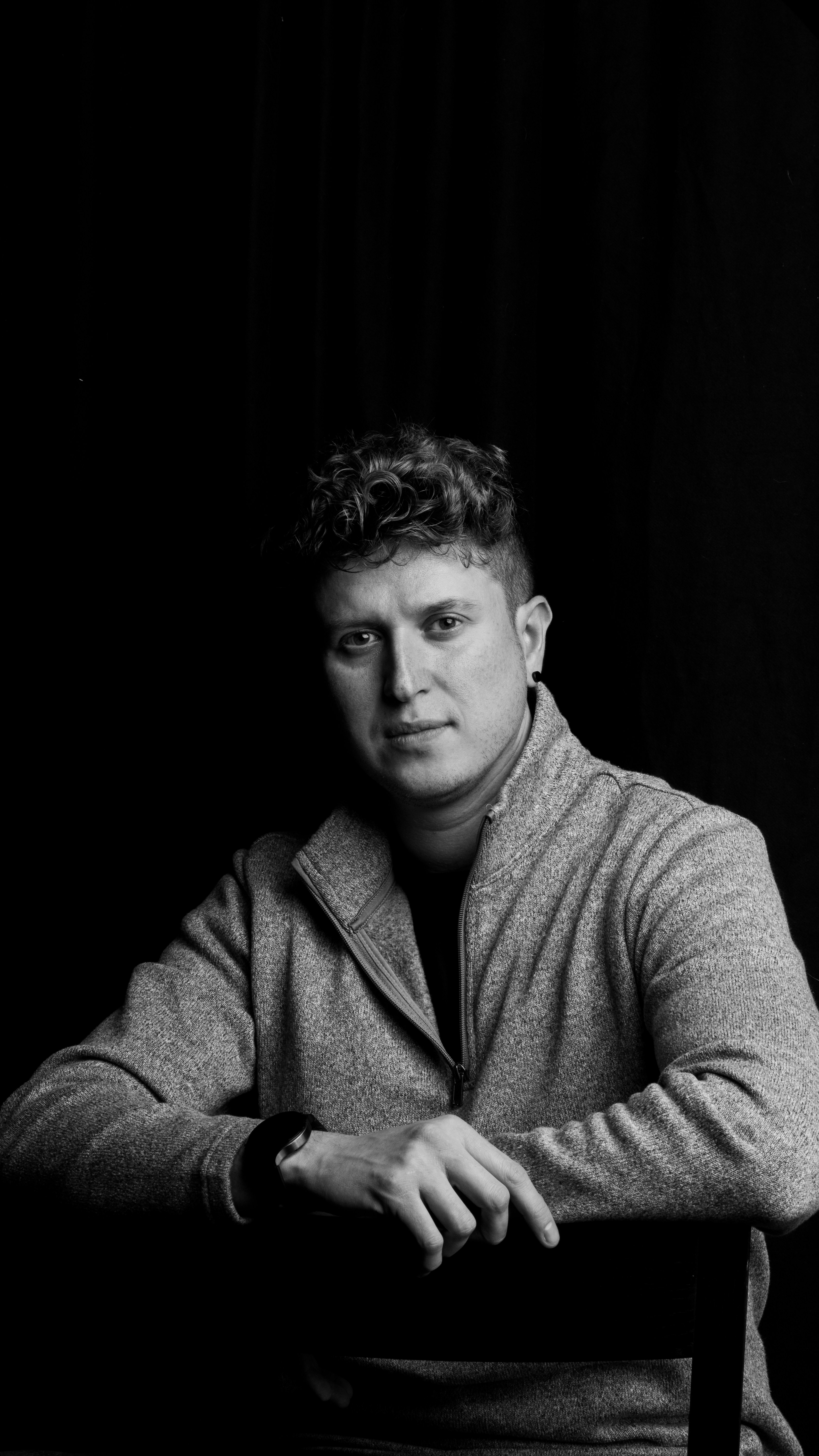 A man with curly hair wearing a grey quarter-zip sweater against a black background