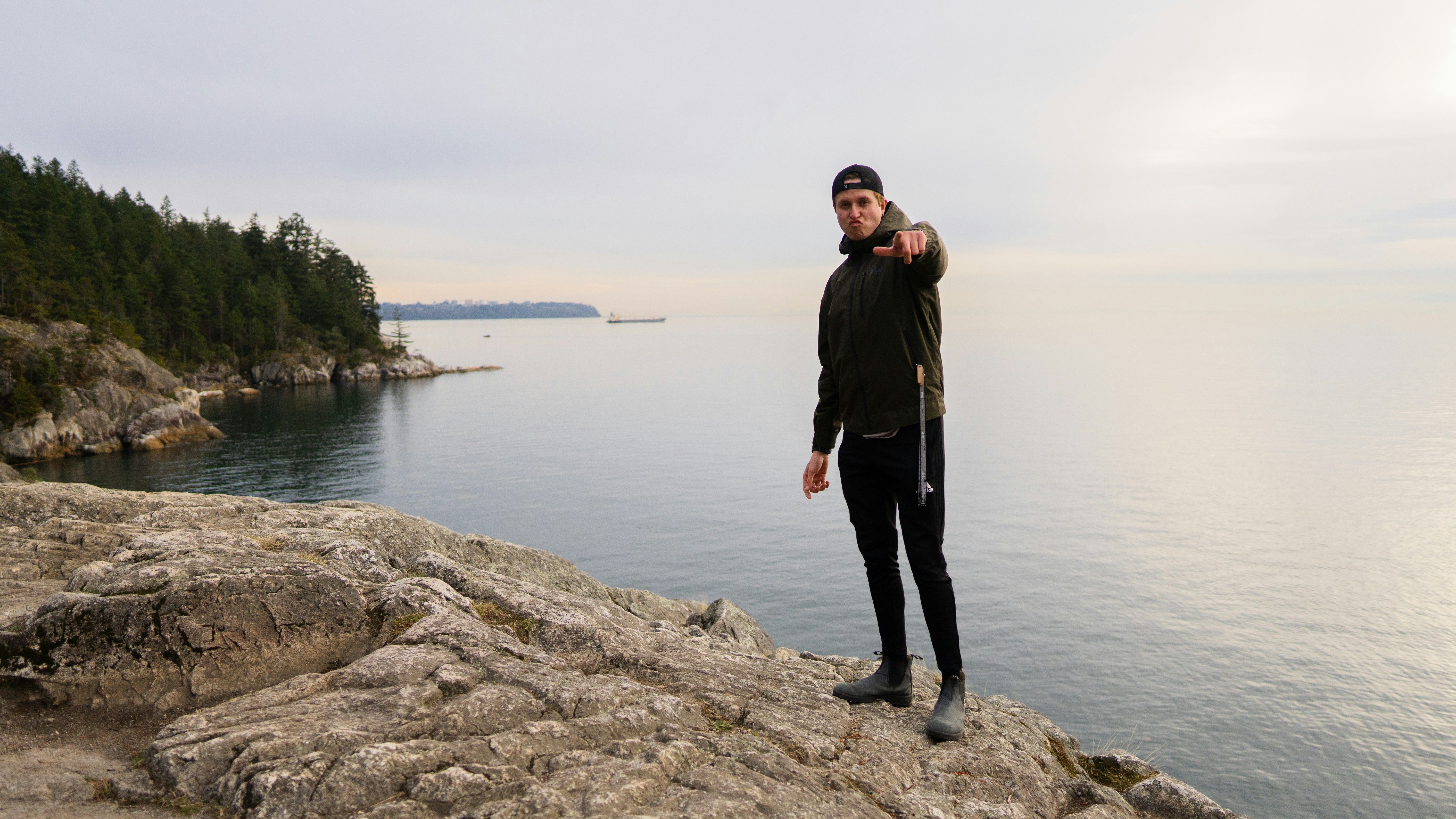 A man in a dark jacket pointing at the camera on a rocky cliff