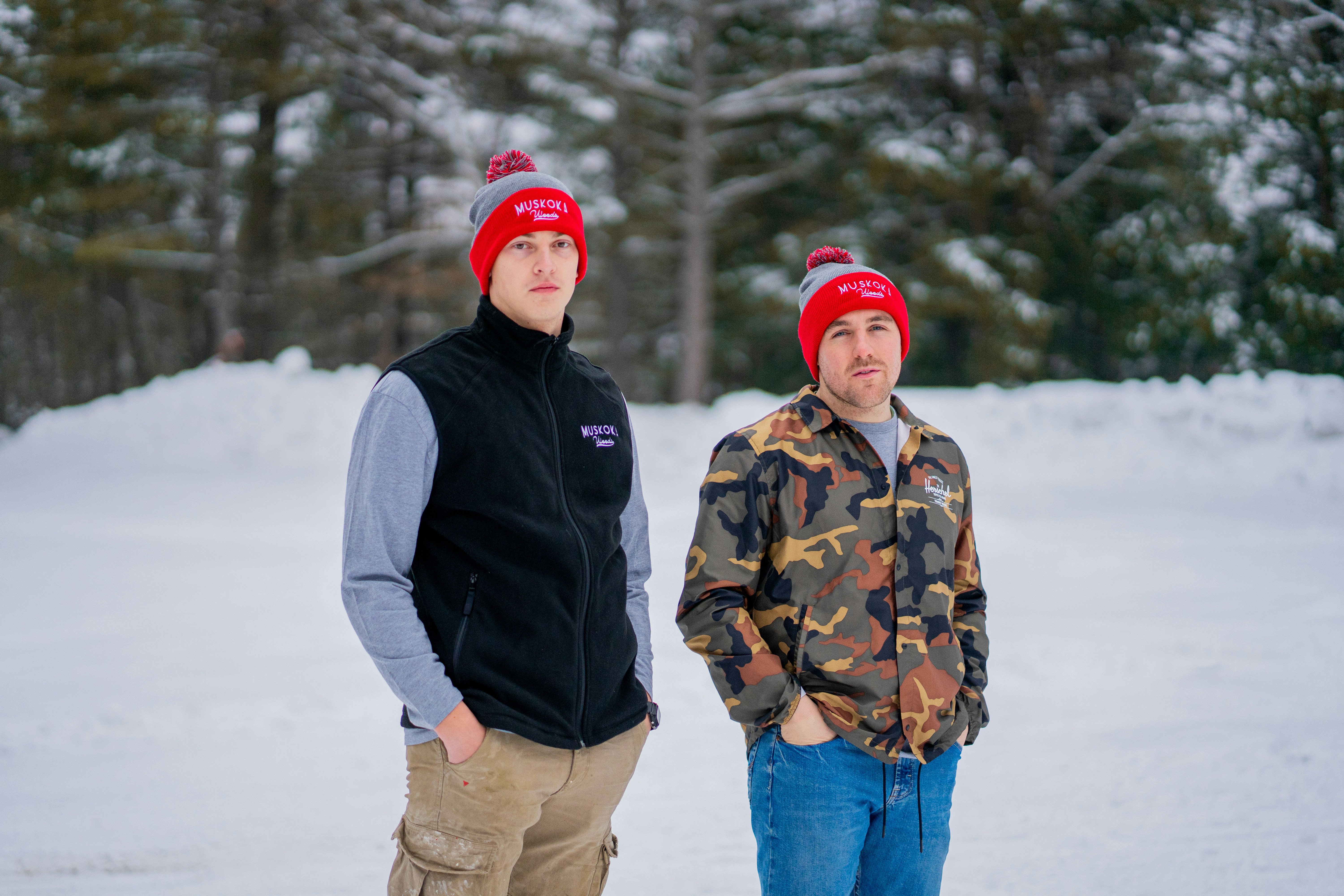 Two men in red and grey beanies standing in a snowy forest clearing