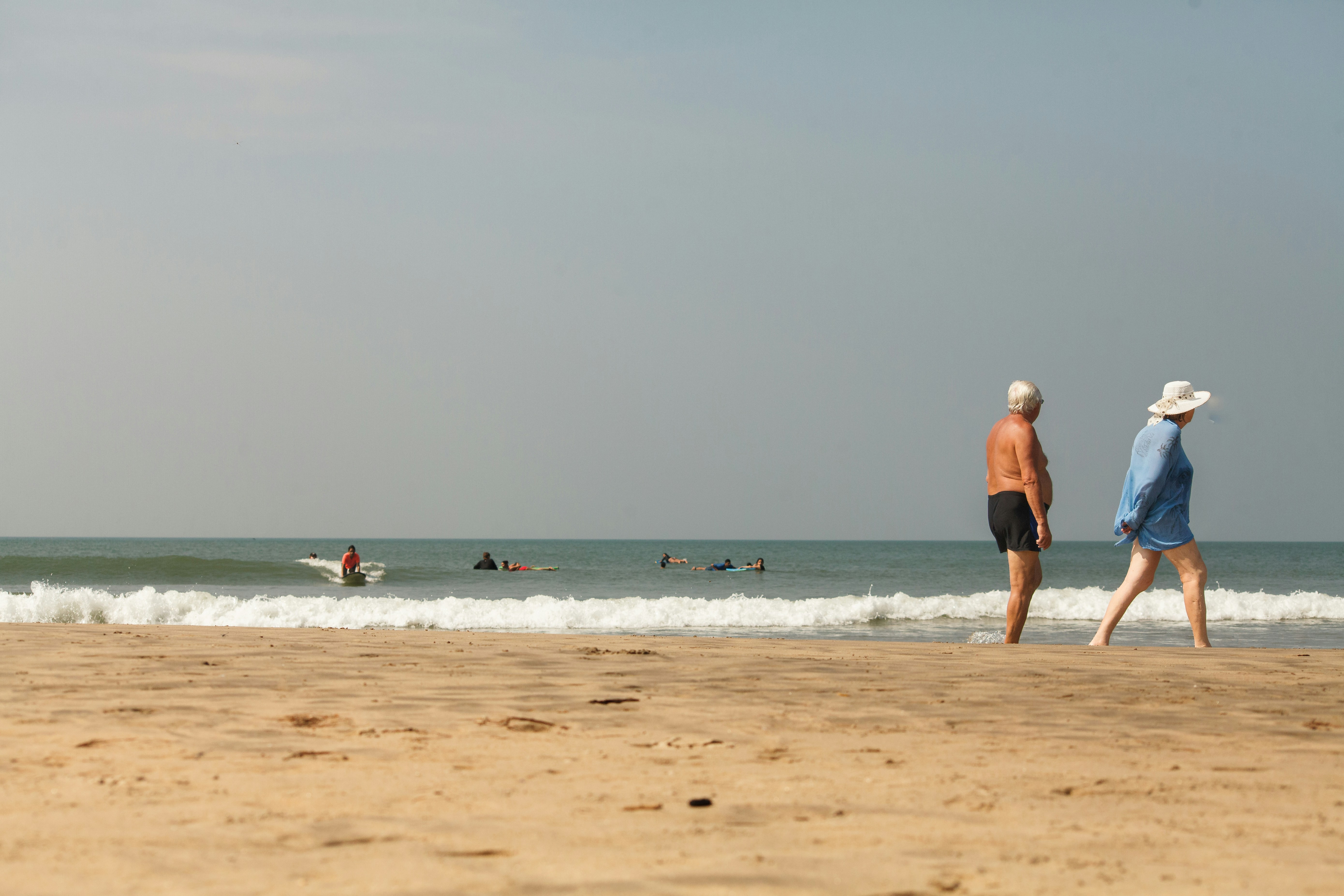 Two adults walking along a sandy beach with surfers in the ocean