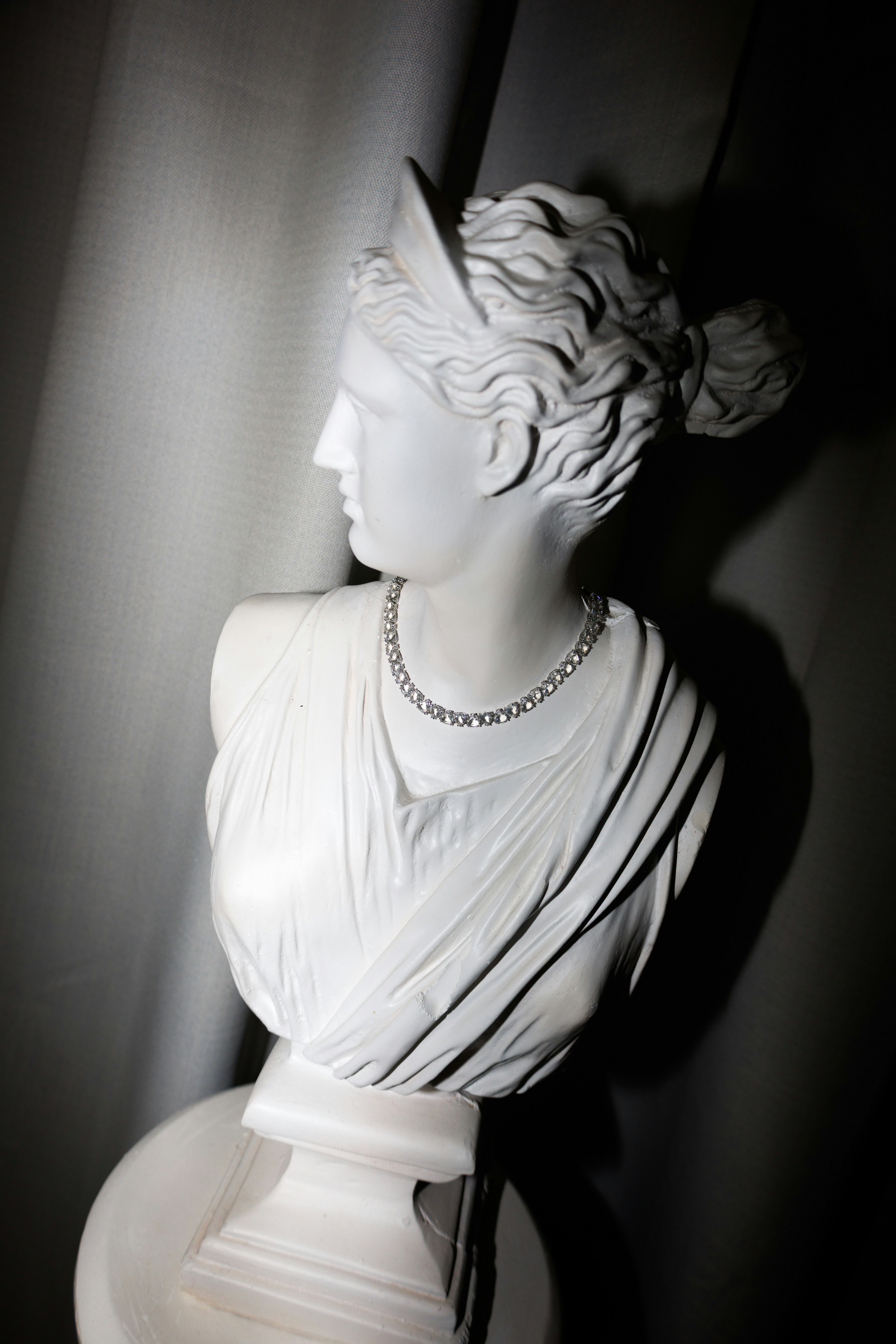 A white classical bust wearing a sparkling diamond necklace against a draped curtain