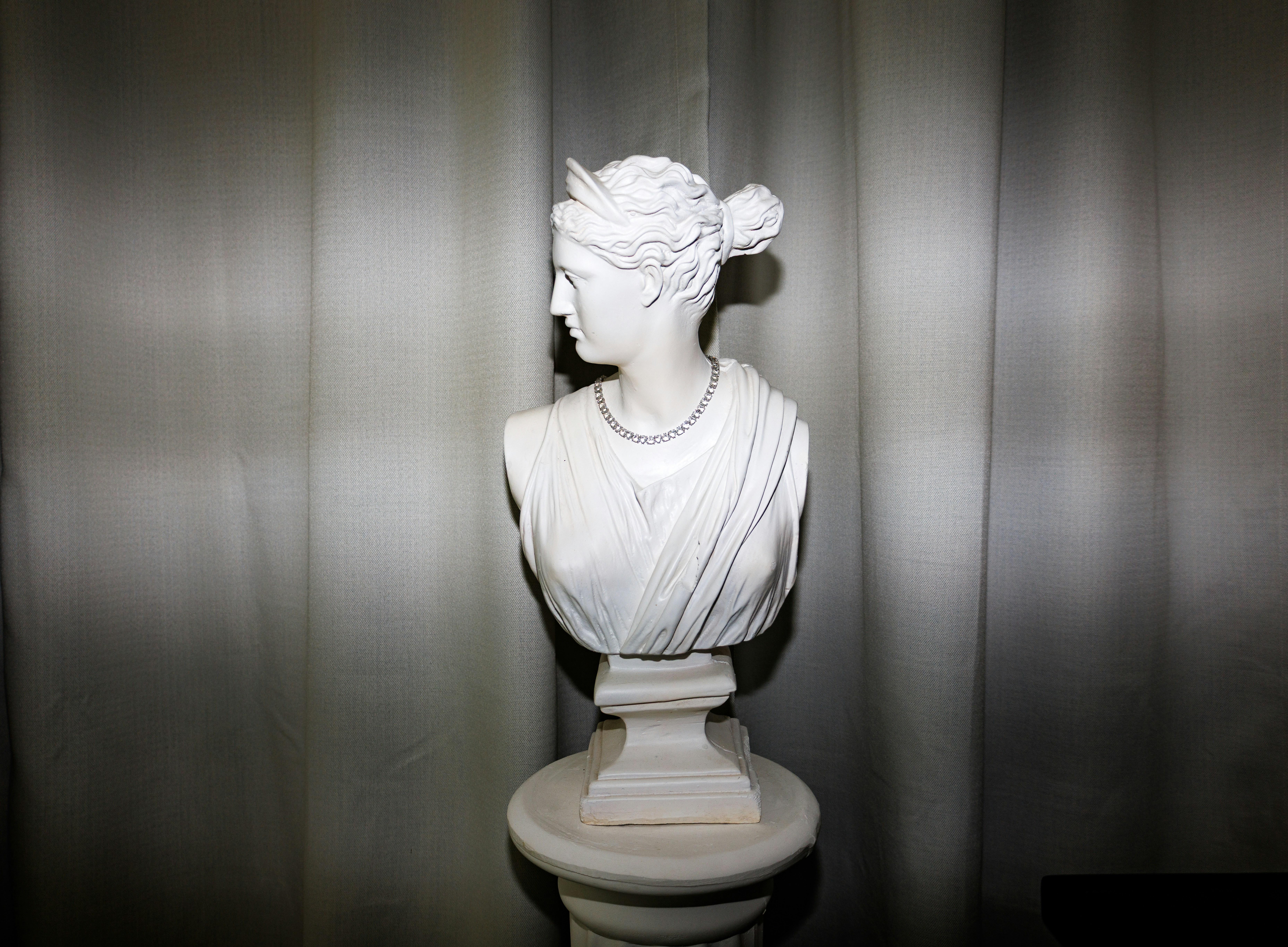 A white plaster bust of a woman wearing a necklace against a curtain