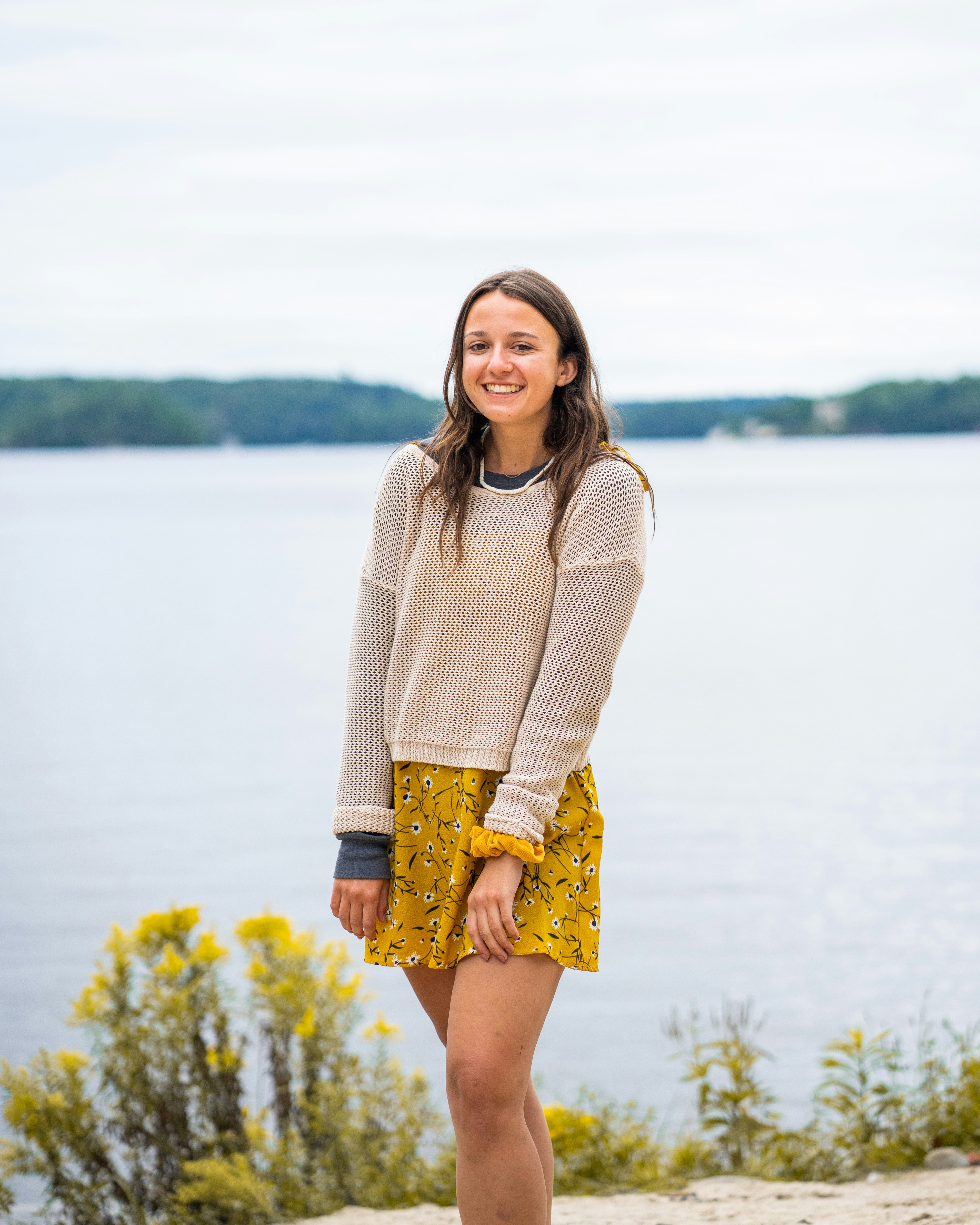 A woman in a beige sweater and yellow floral dress by a lake