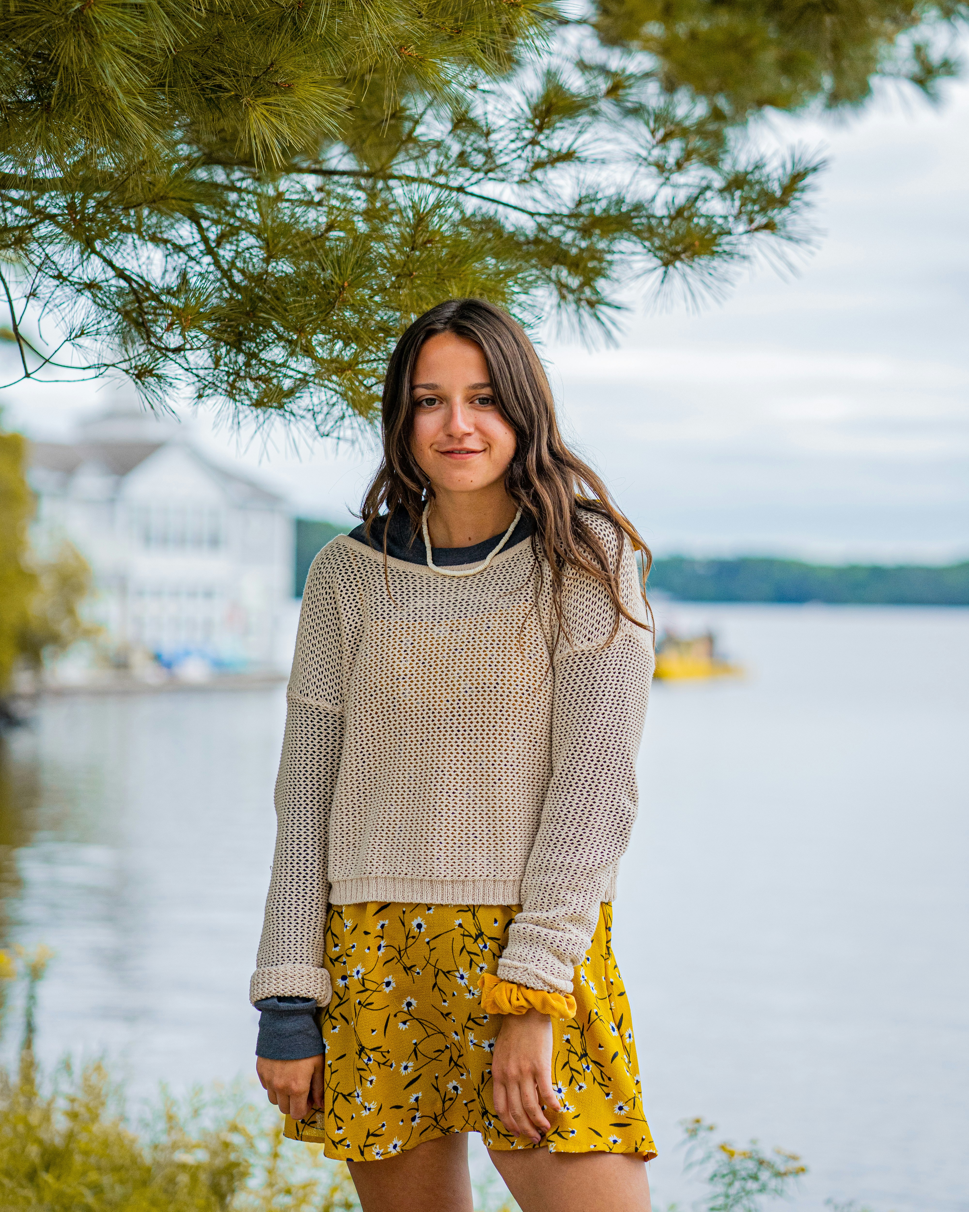 A woman in a beige knit sweater and yellow floral dress by a lake