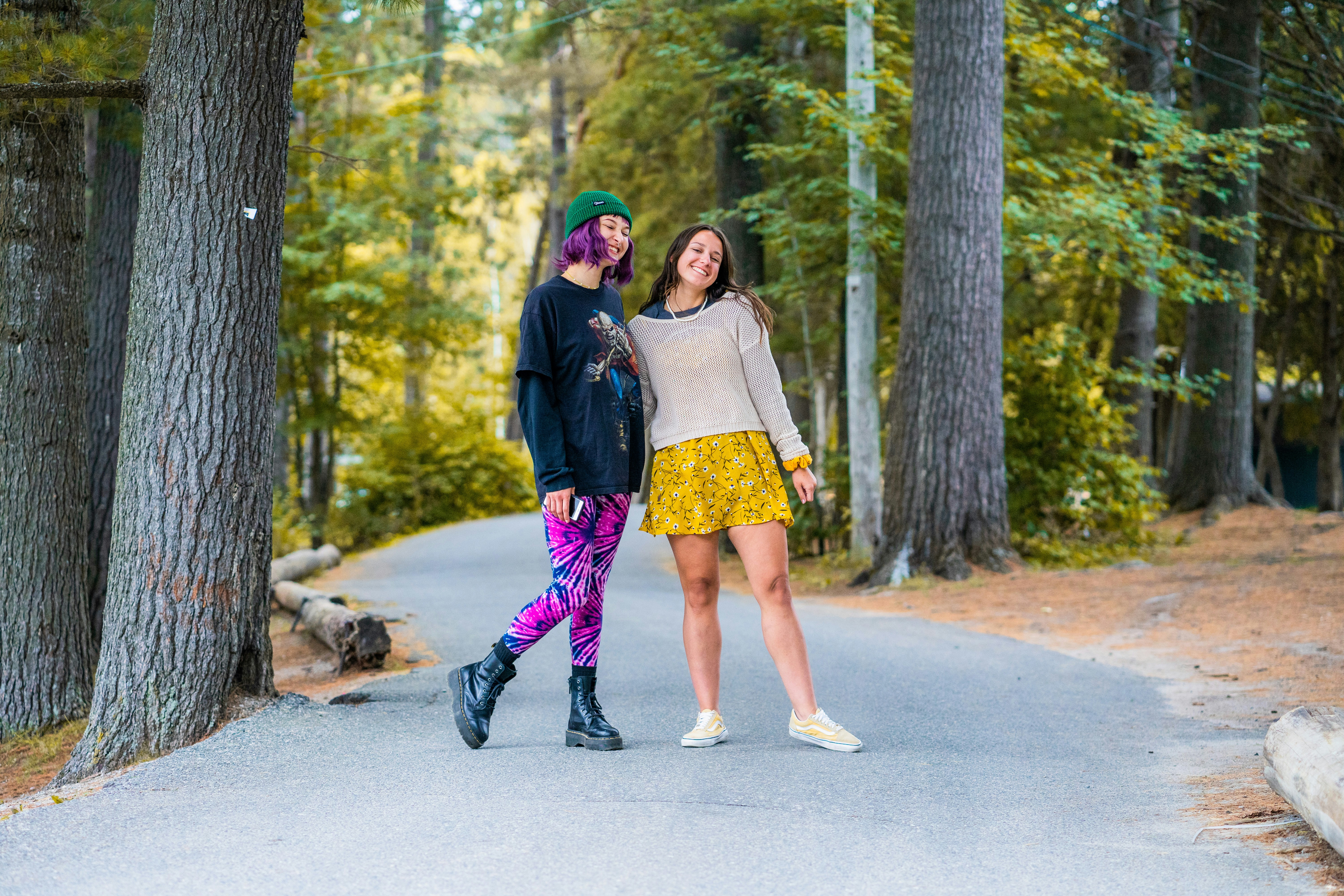 Two young women smiling on a paved path through a forest of tall trees