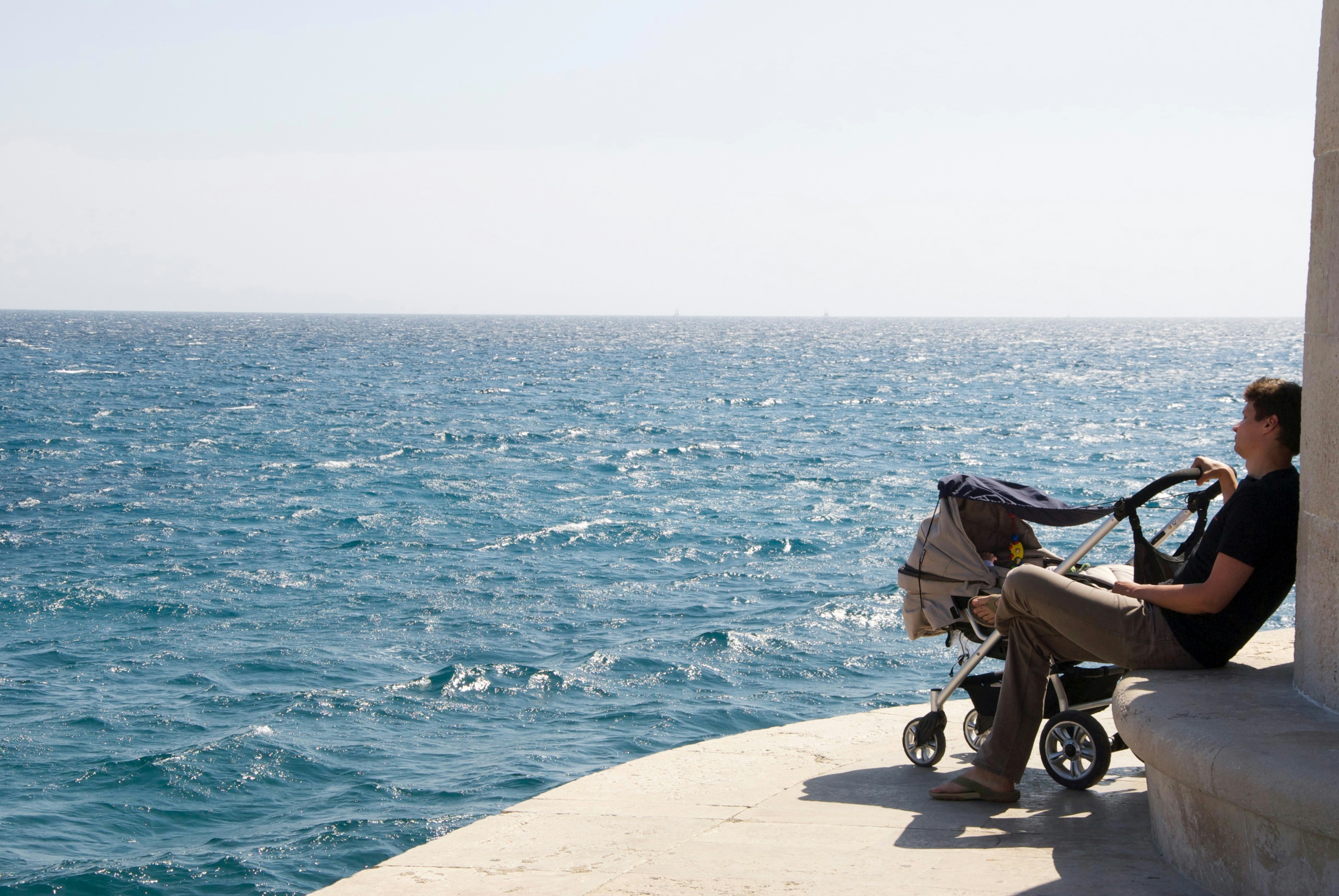 Man sitting on a seawall ledge beside a lightweight stroller, looking out over a choppy blue sea