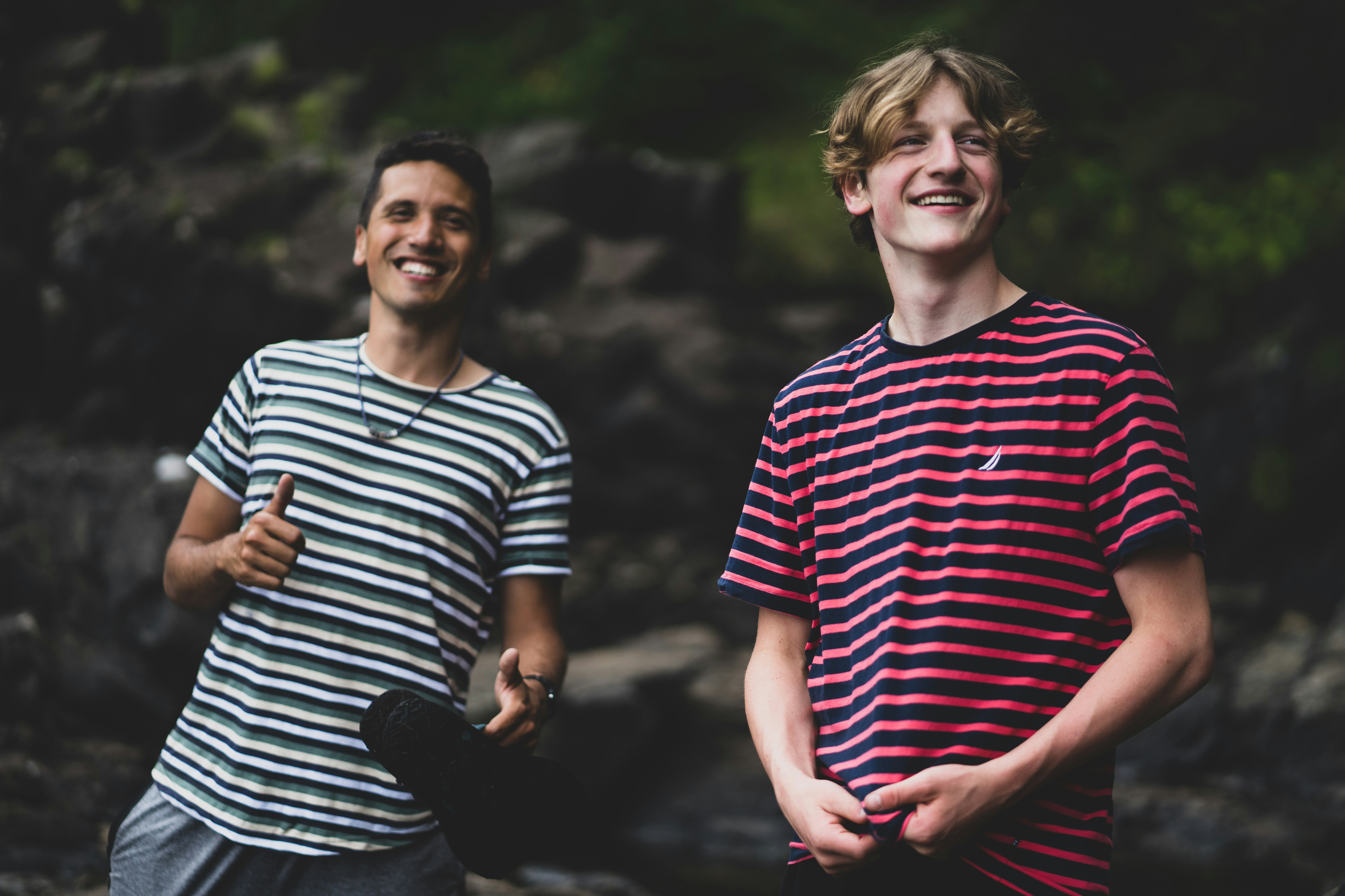 Two men in striped t-shirts smiling outdoors with one giving a thumbs up