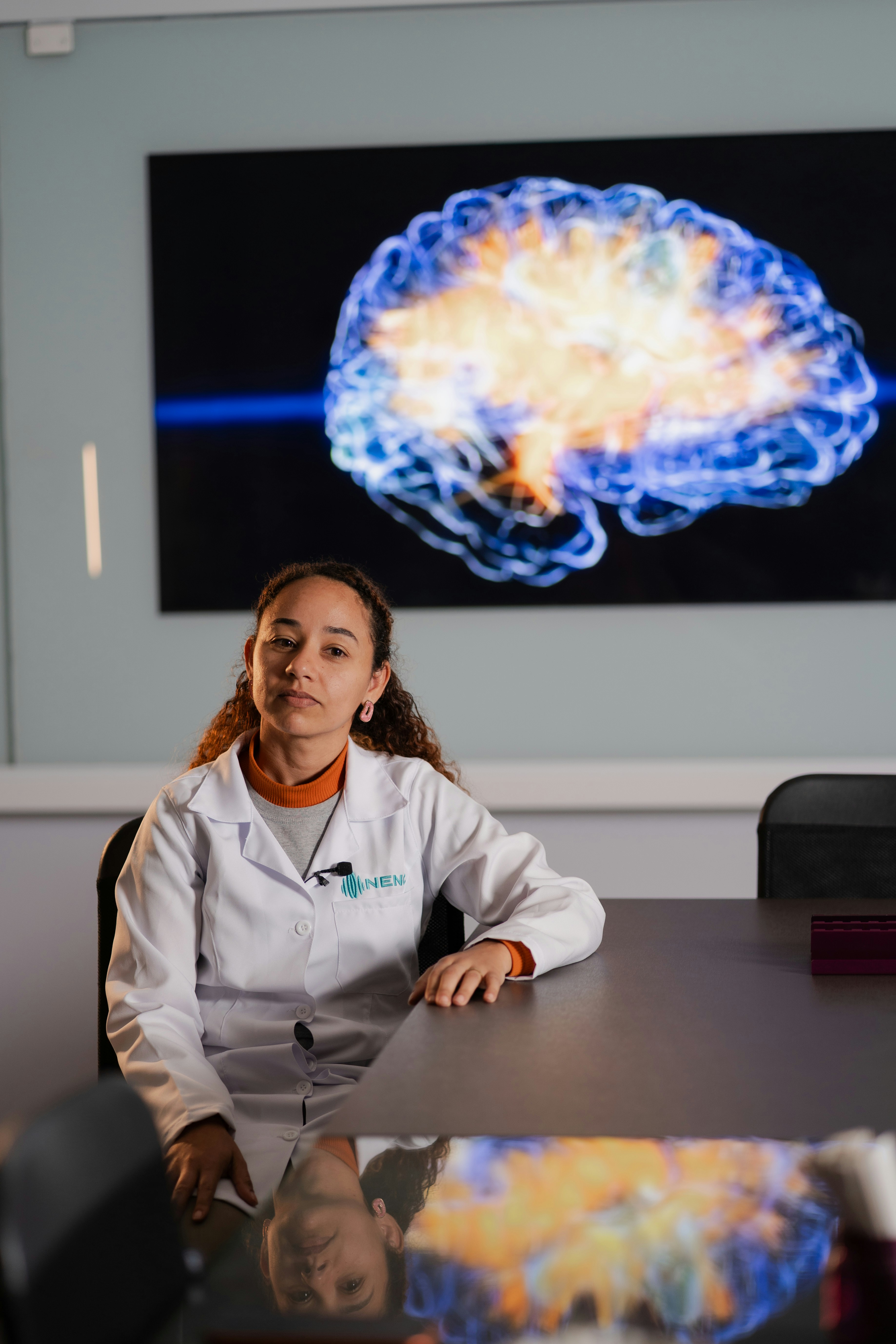 A woman in a white lab coat sitting at a table before a brain scan