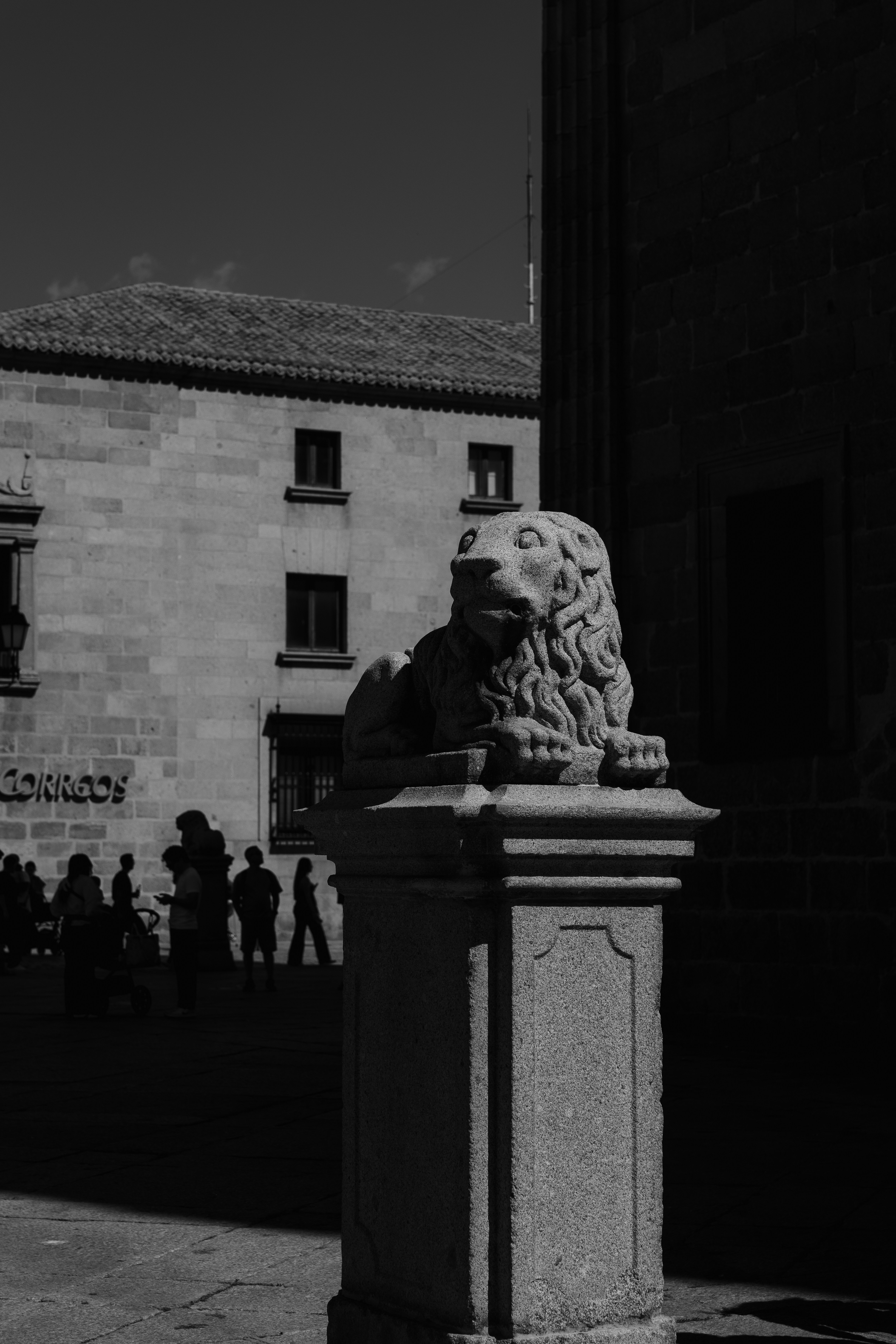 A stone lion statue on a pedestal in a plaza with people walking