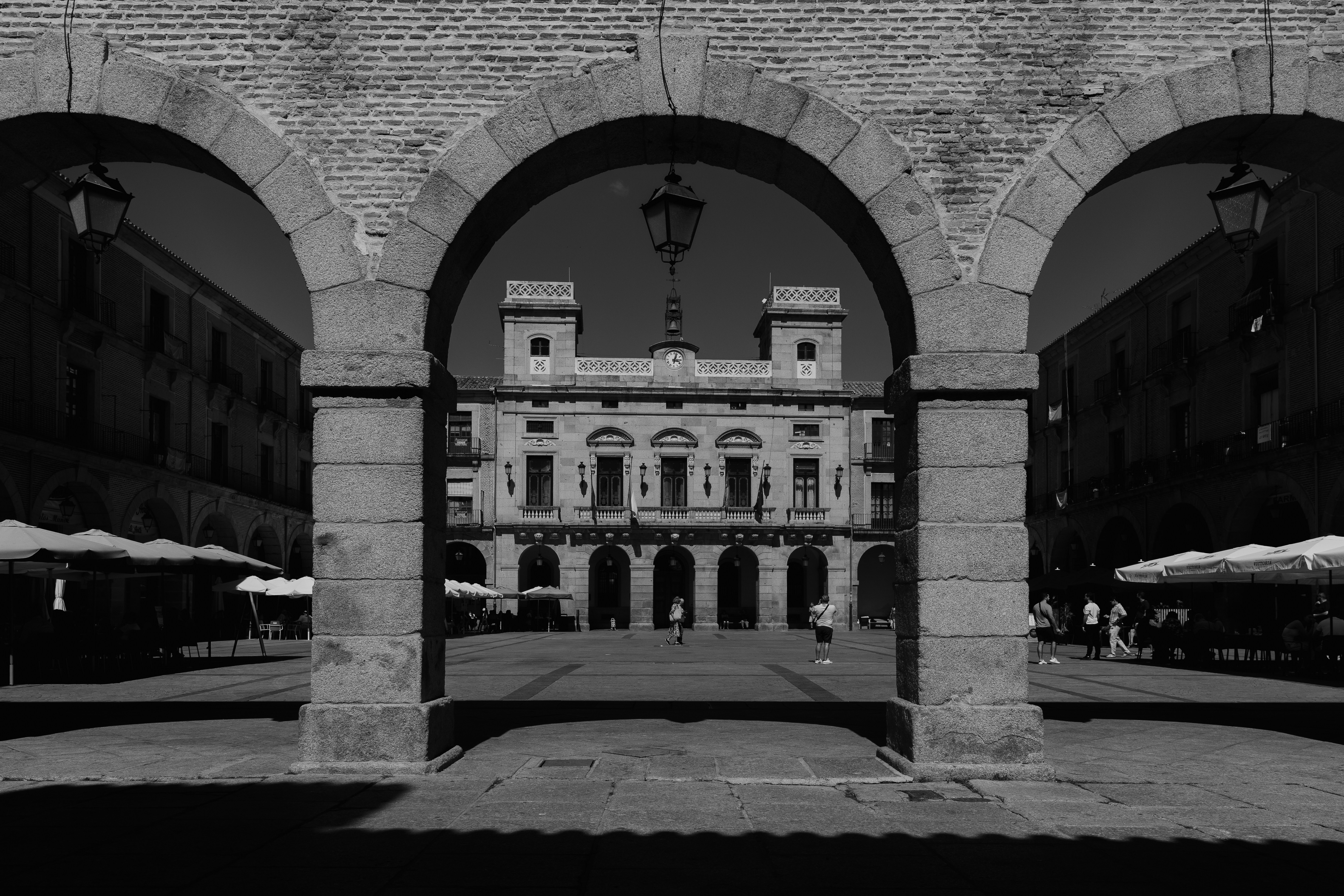 A stone archway framing a historic town square with people walking in the distance