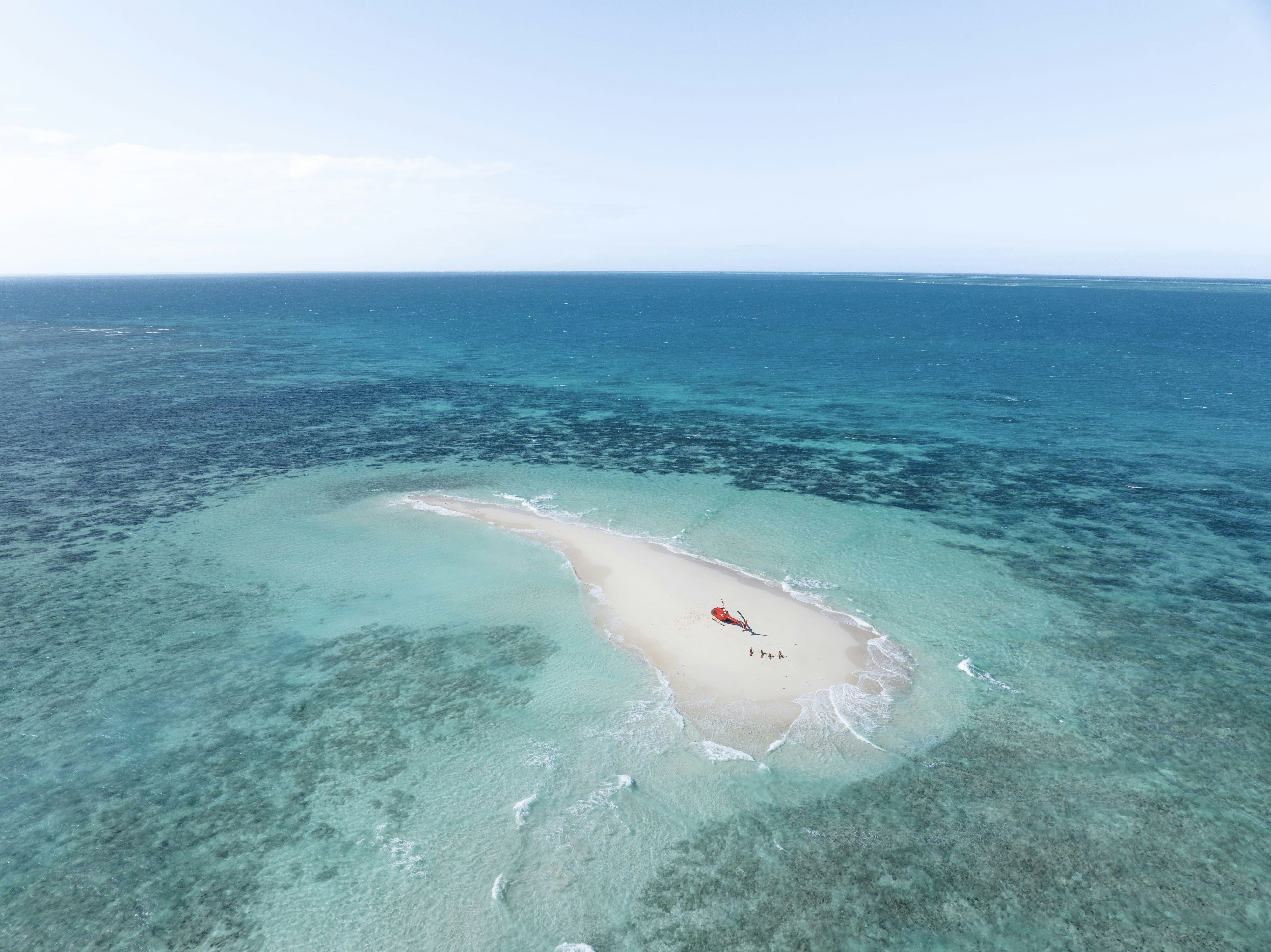 A red helicopter on a small white sandbar surrounded by turquoise ocean water