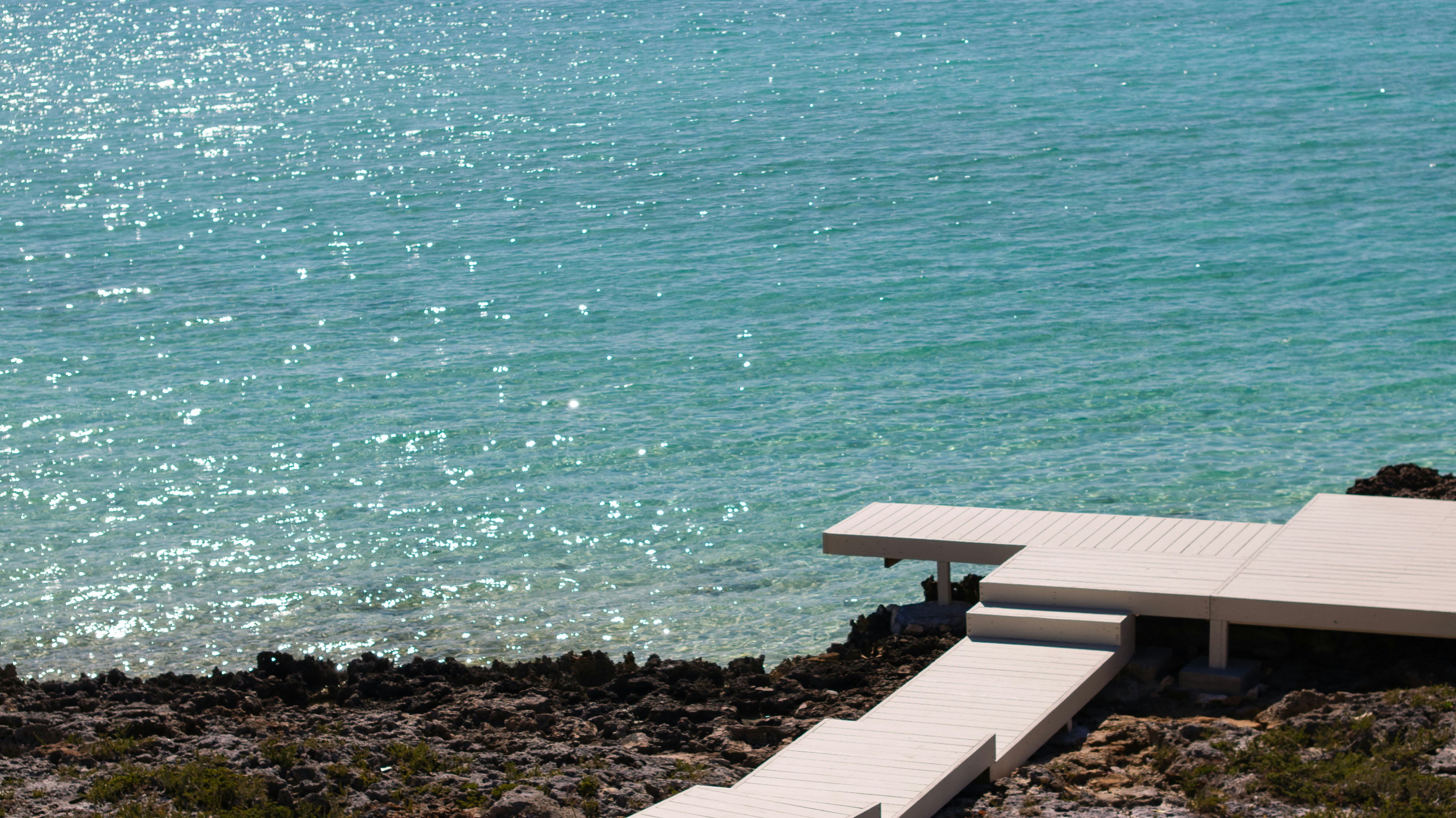 A white wooden deck extending over clear turquoise water from a rocky shore