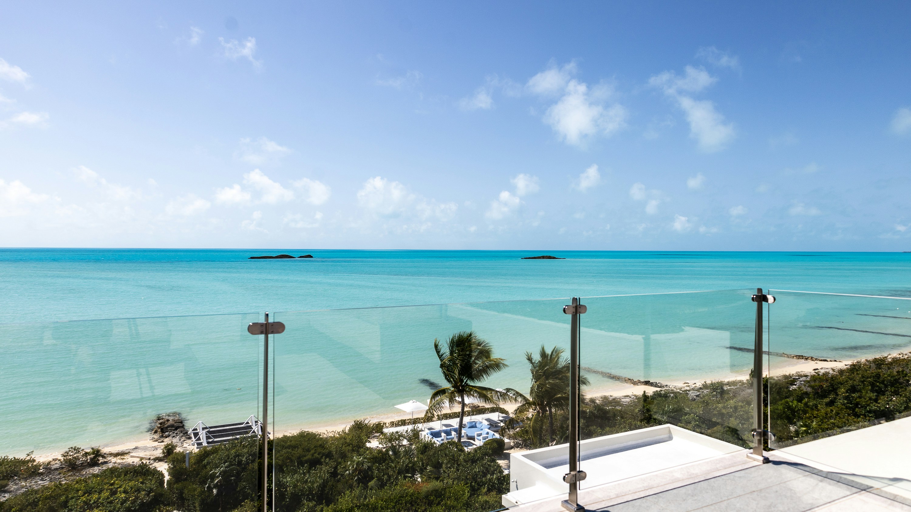 A glass balcony railing overlooking a turquoise sea with a sandy beach and palm trees