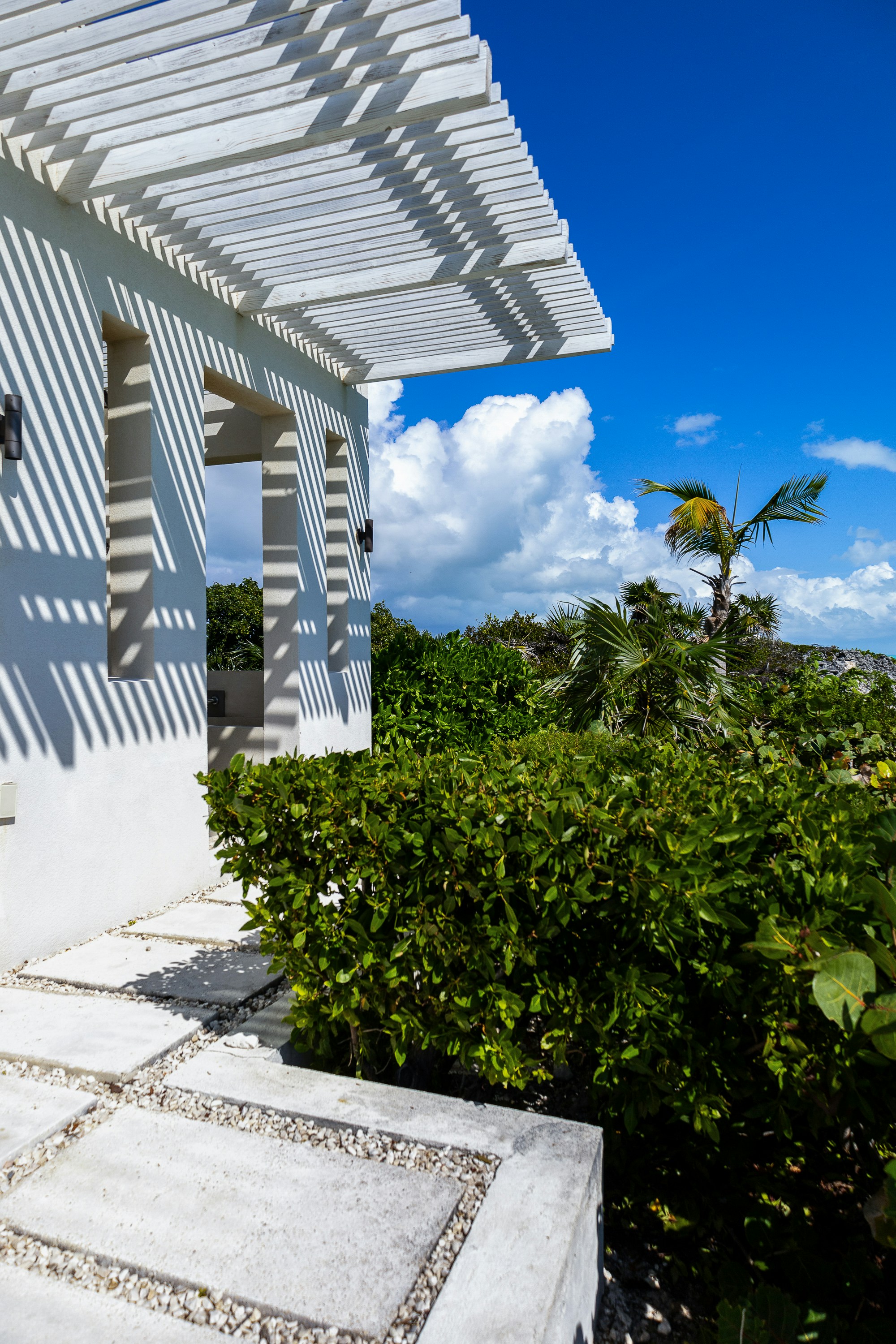 A white building with a wooden pergola casting shadows over a garden path