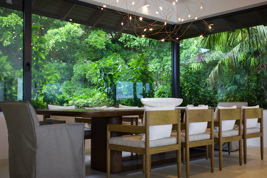 Timber dining table beside floor-to-ceiling windows and tropical garden