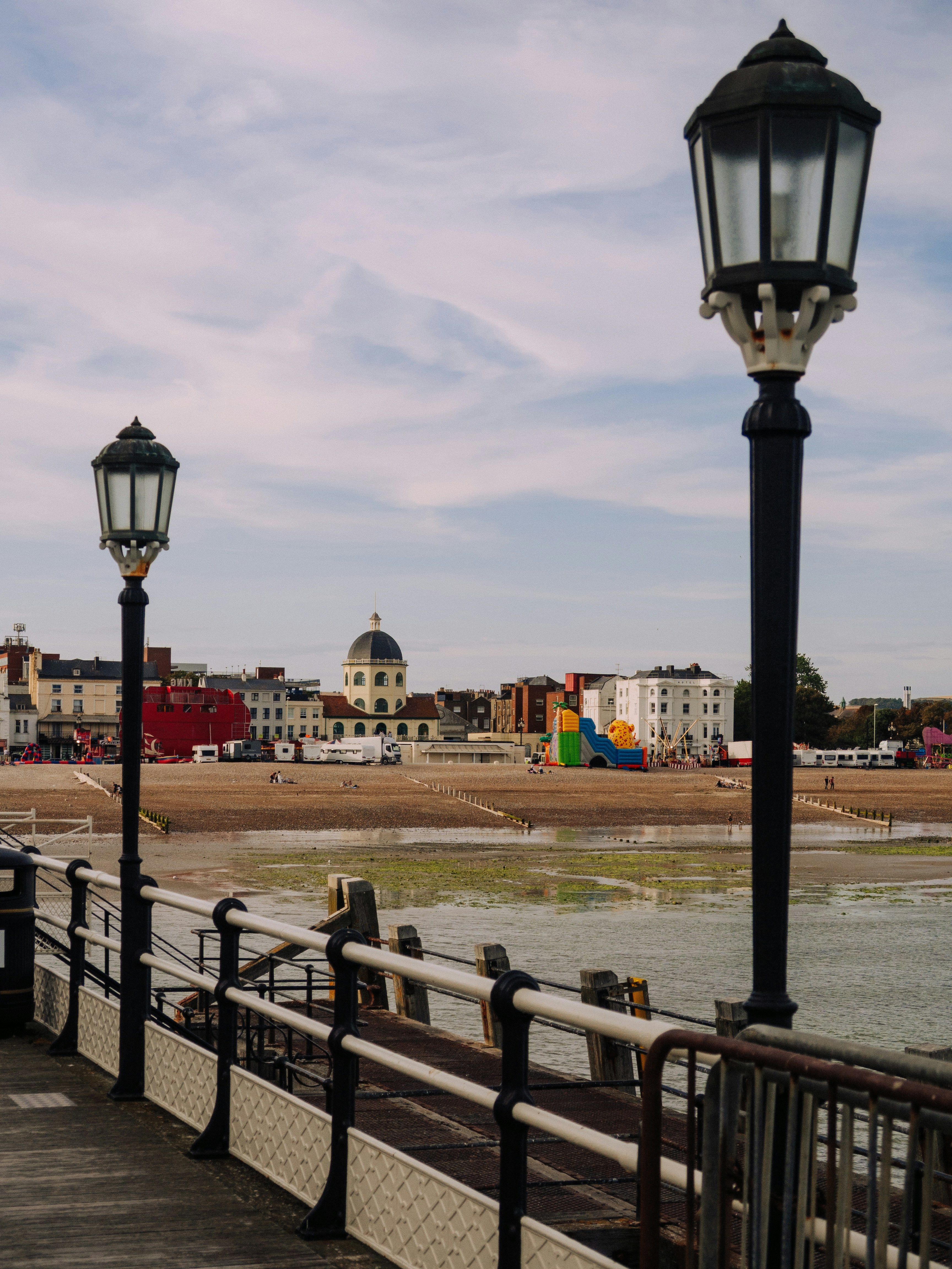 Two ornate black lampposts on a wooden pier overlooking a pebble beach and town