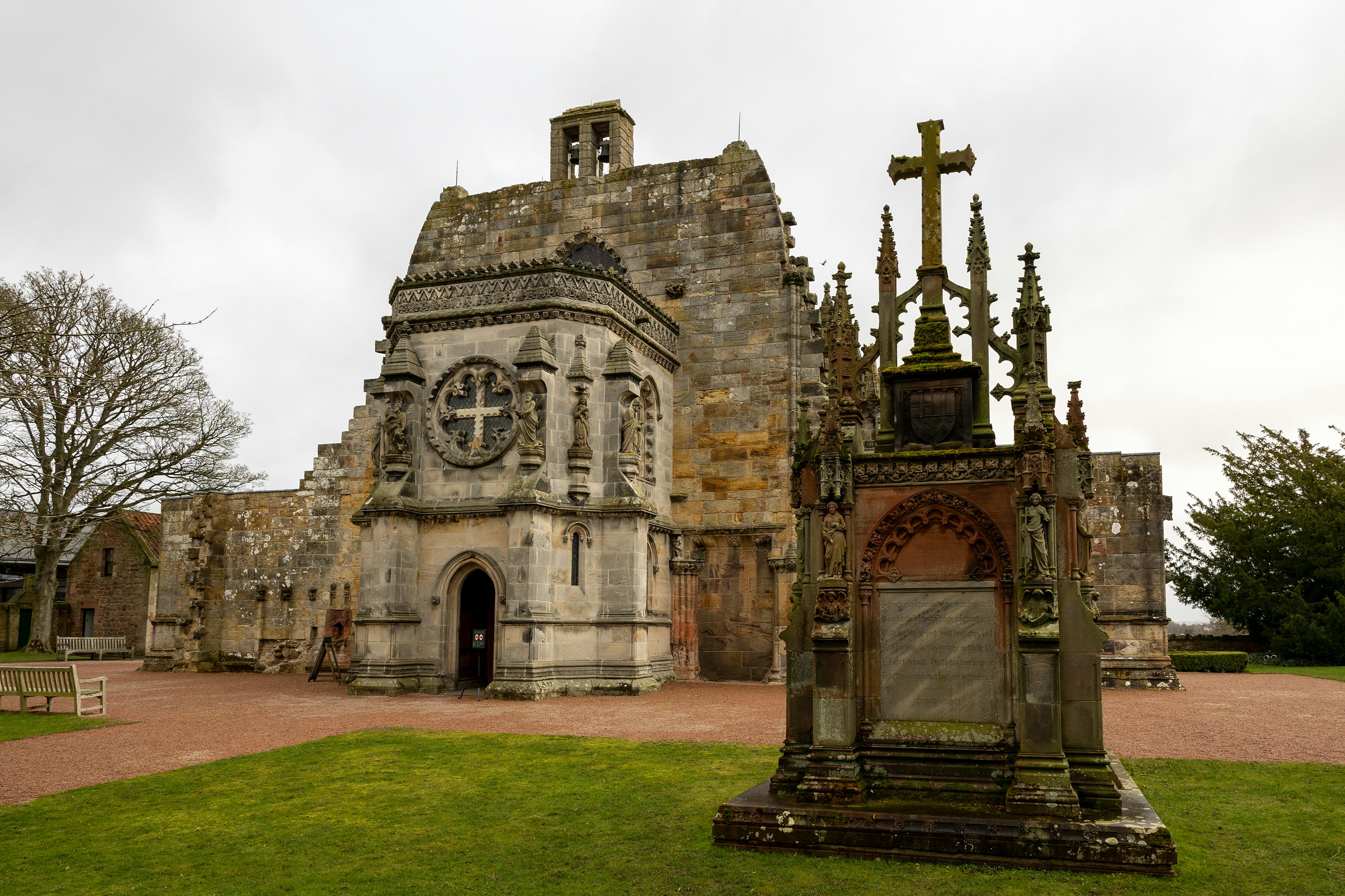 The stone Rosslyn Chapel with an ornate cross monument in the foreground
