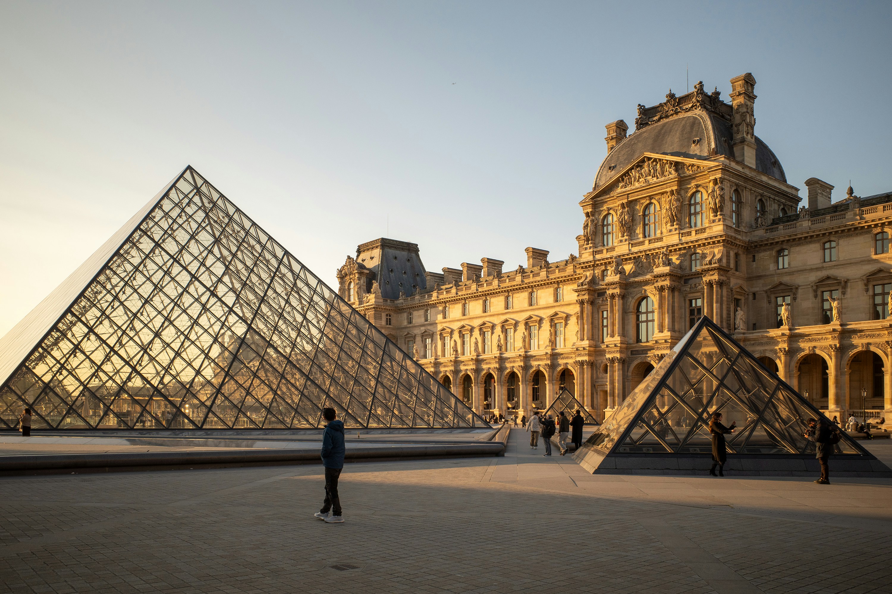 People walking near the glass Louvre Pyramid in Paris during the golden hour