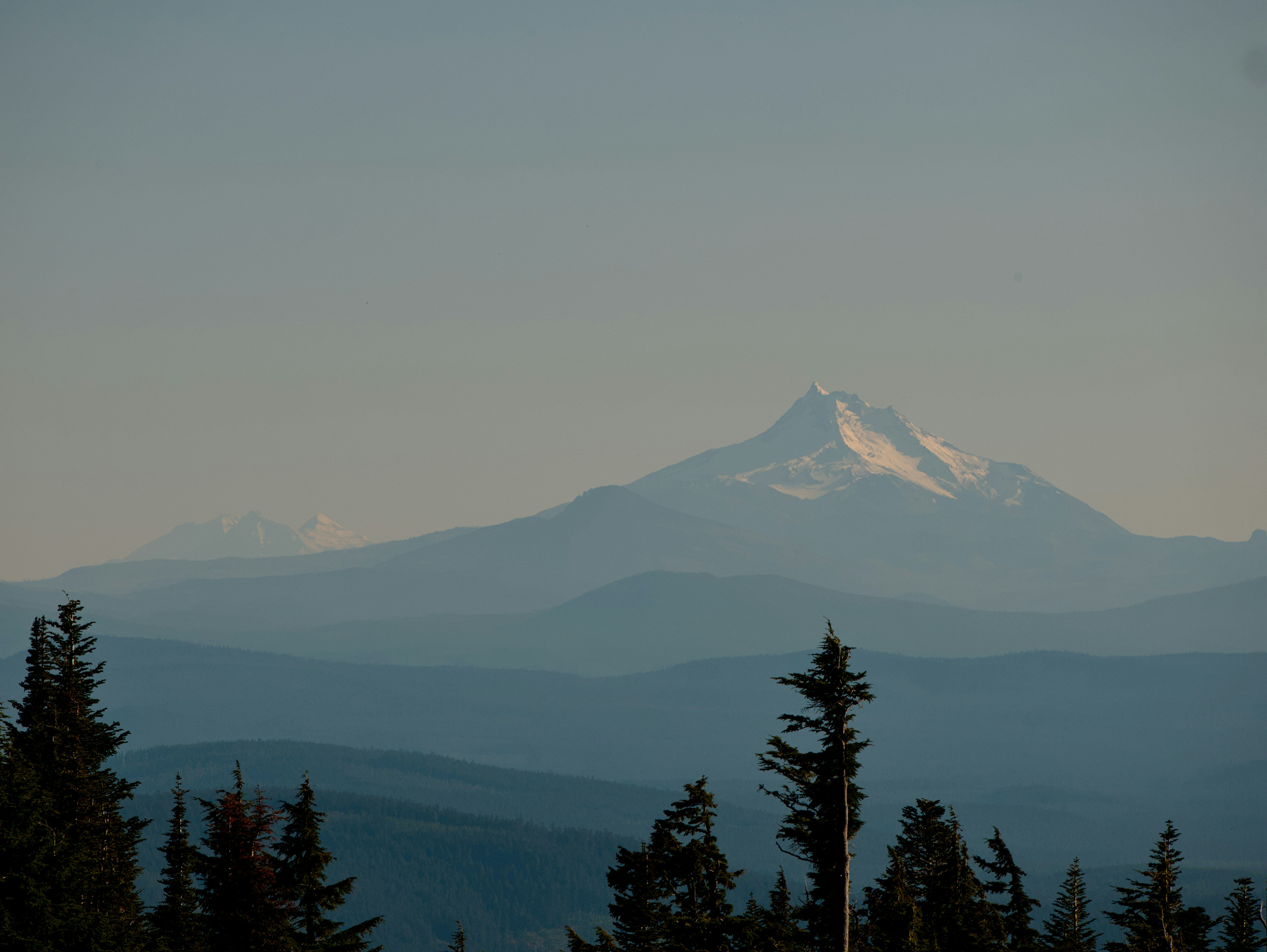 A snow-capped mountain peak rising above layers of hazy blue forested hills