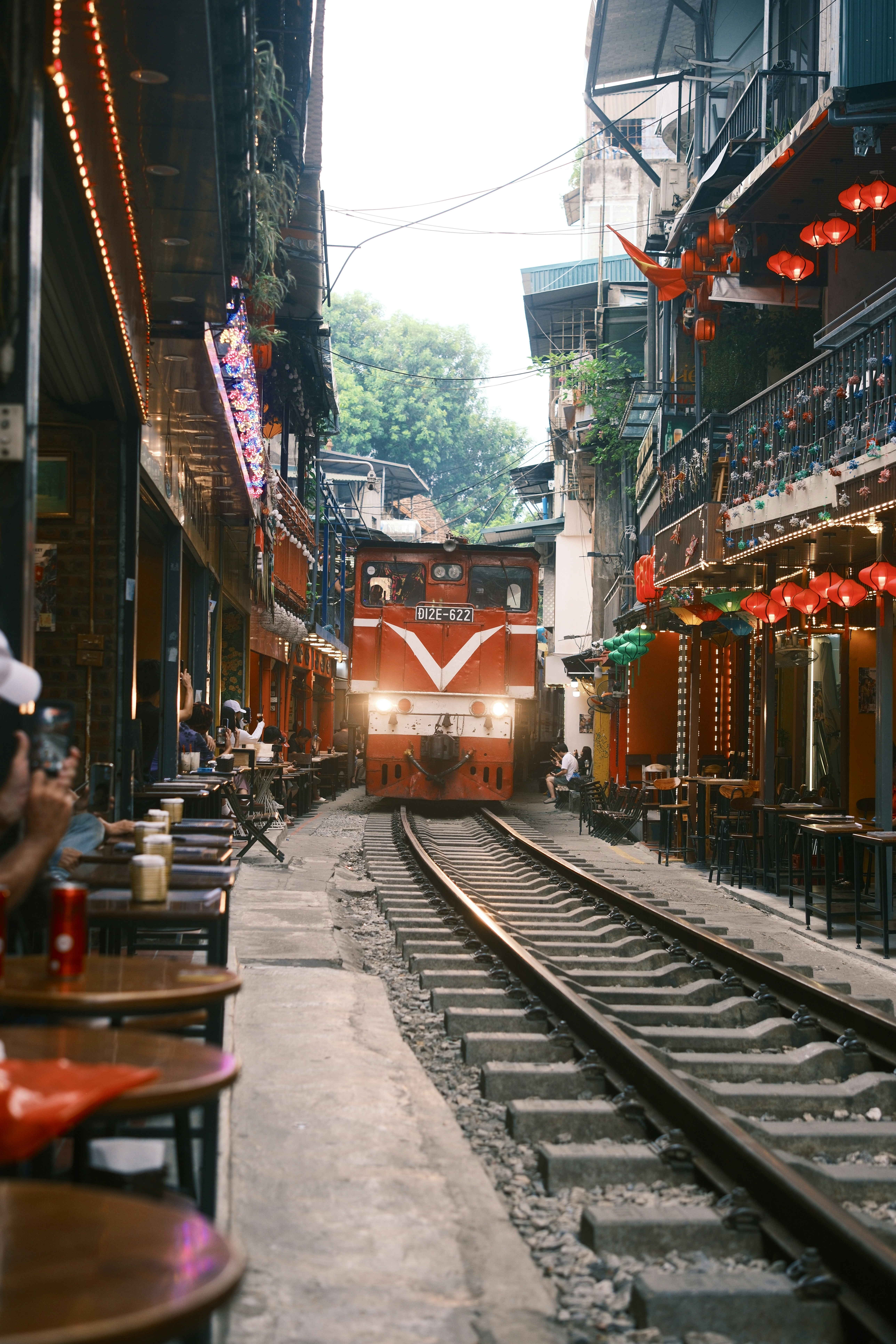 A red train moving along tracks through a narrow street lined with cafes in Hanoi