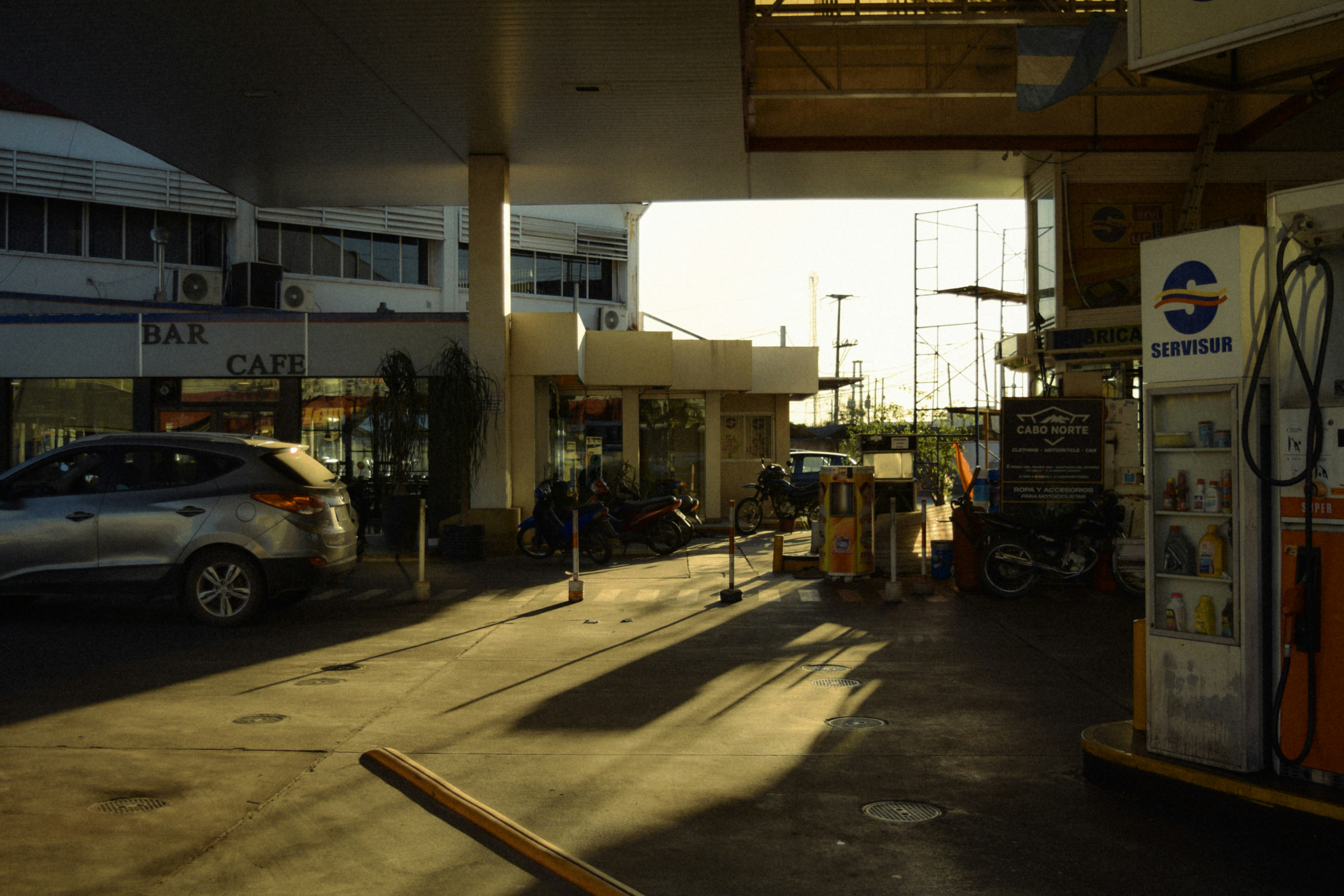 Una gasolinera con un coche plateado y motos bajo una marquesina bajo el sol bajo