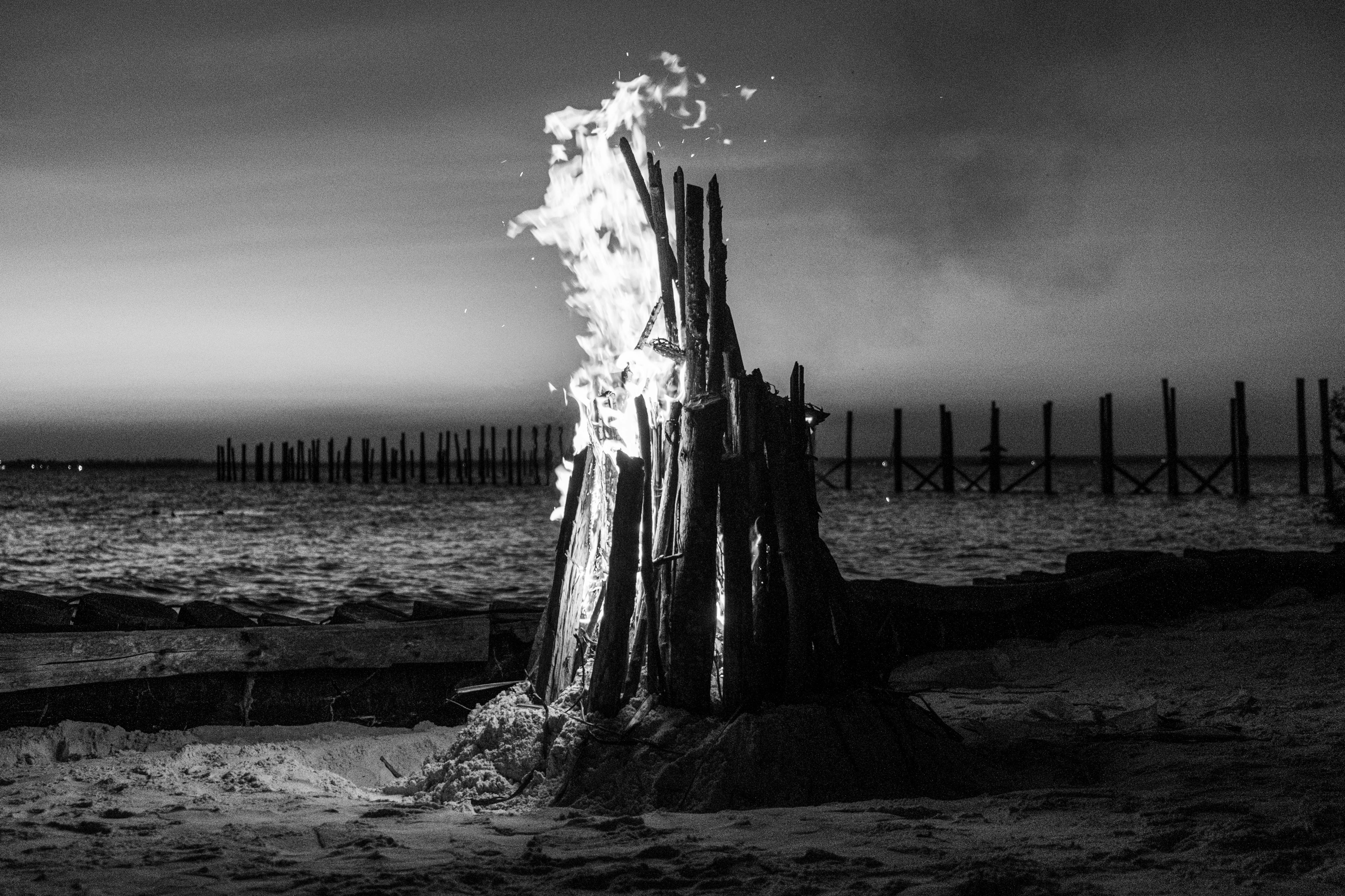 A bonfire burning on a sandy beach at night with wooden pilings offshore
