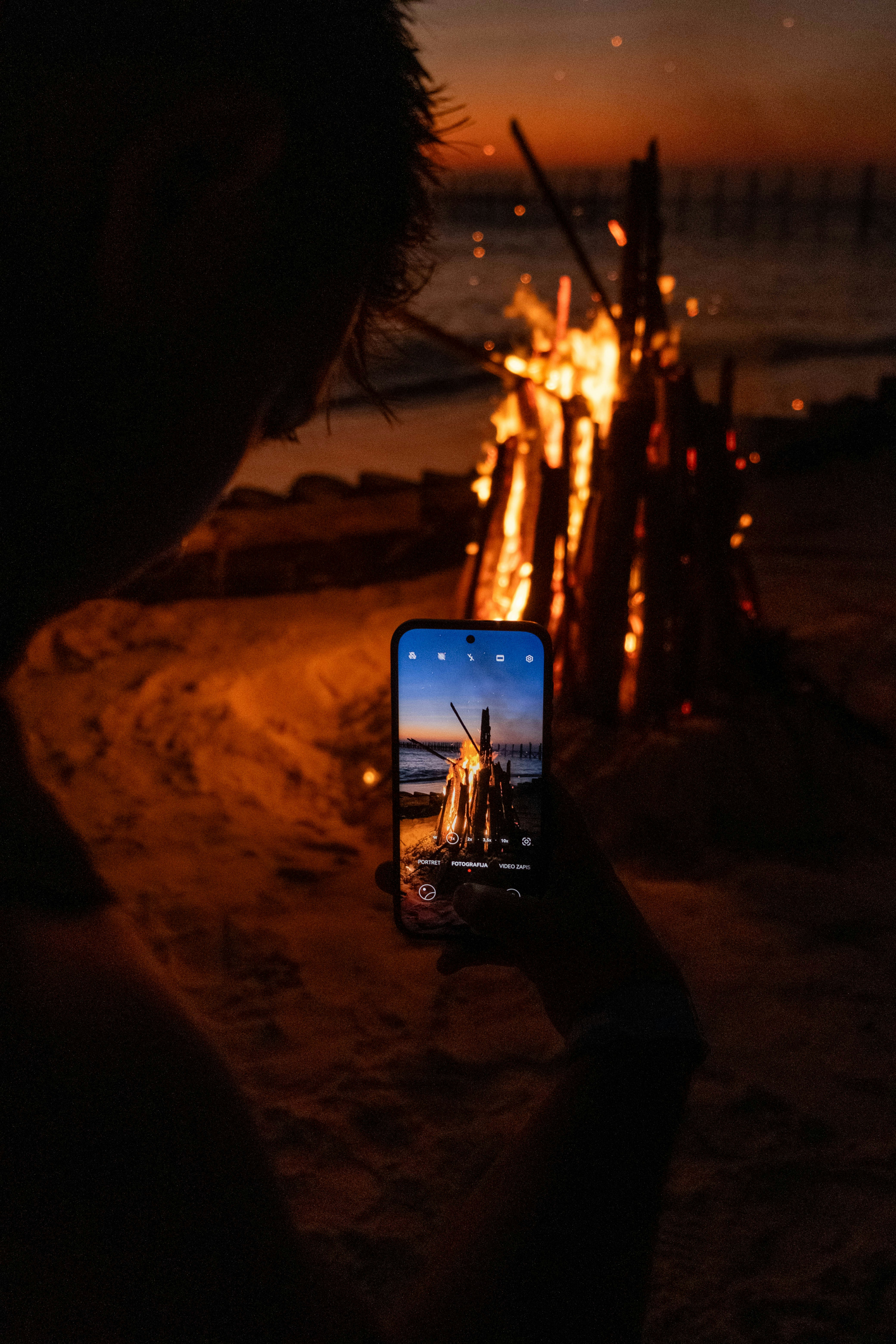 Une personne tenant un smartphone pour photographier un feu de joie sur une plage de sable