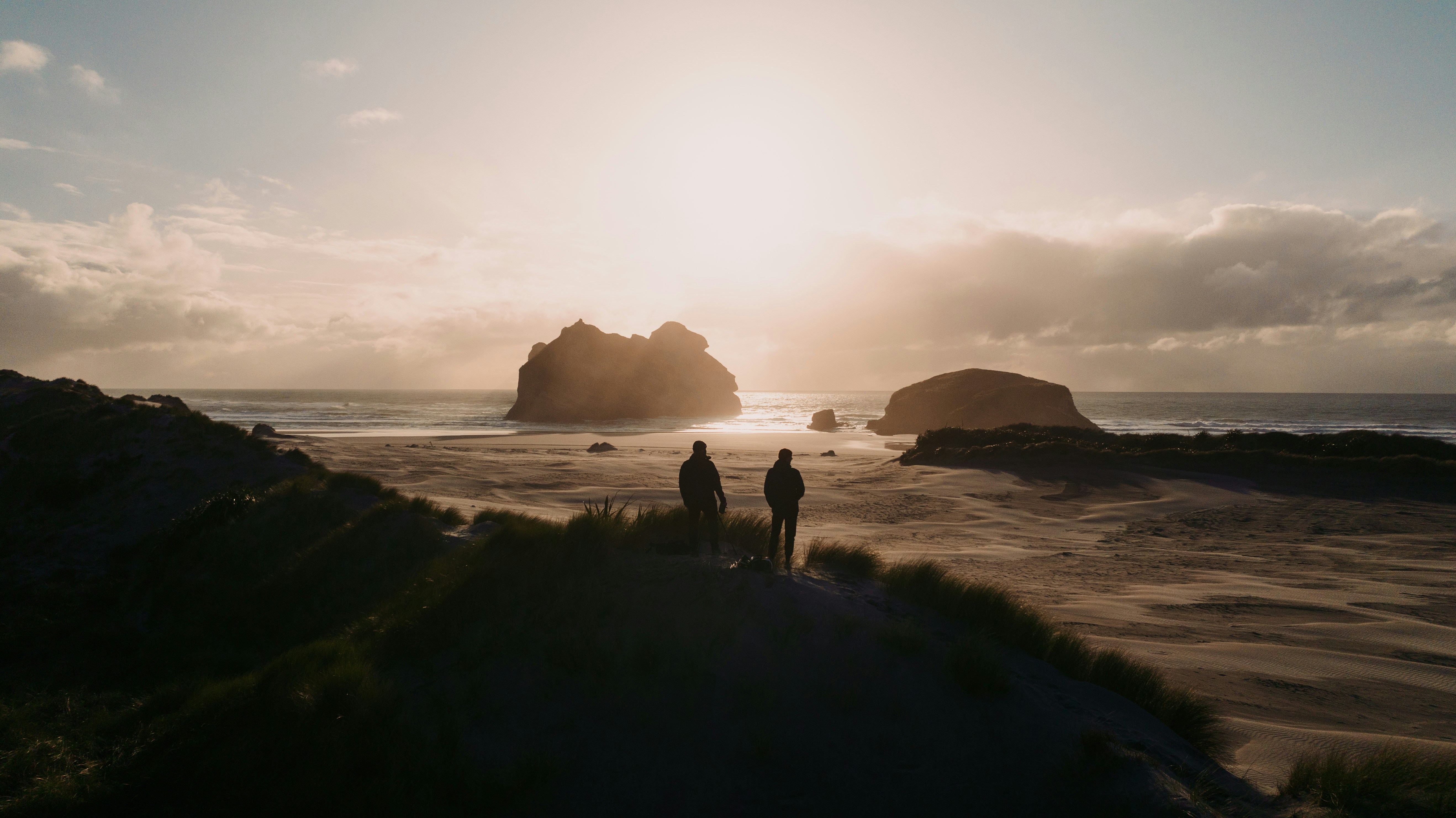 Two people in silhouette on a grassy dune overlooking a beach at low sun