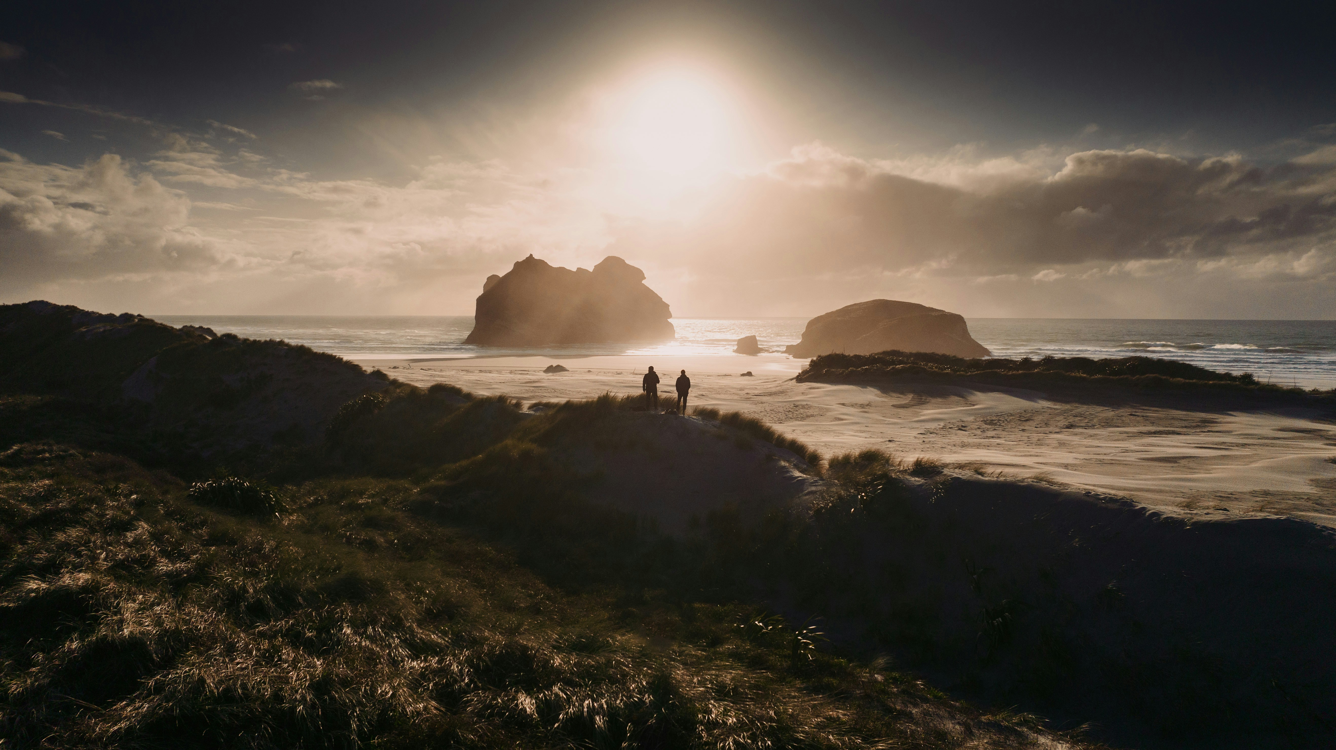 Two people on a grassy dune overlooking a sandy beach at low sun