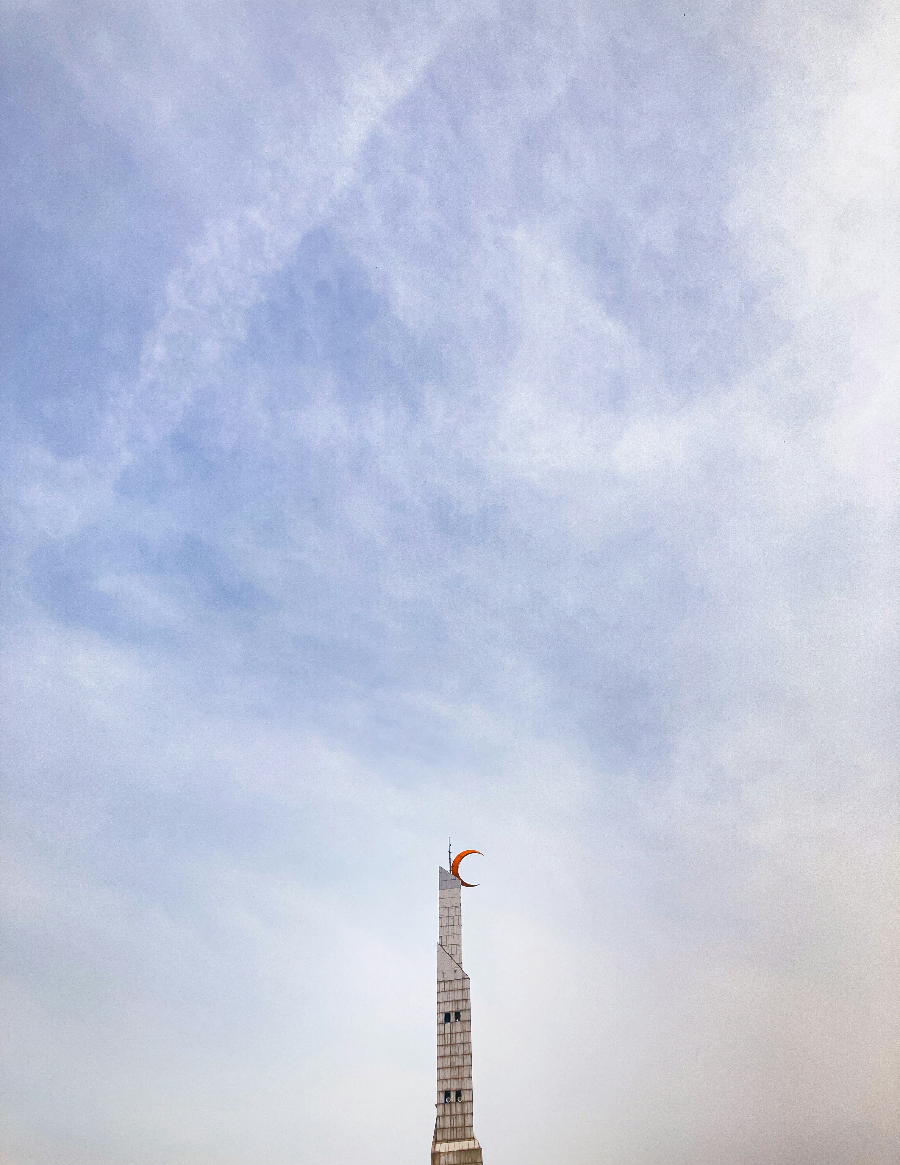 A tall white minaret topped with an orange crescent moon against a cloudy sky