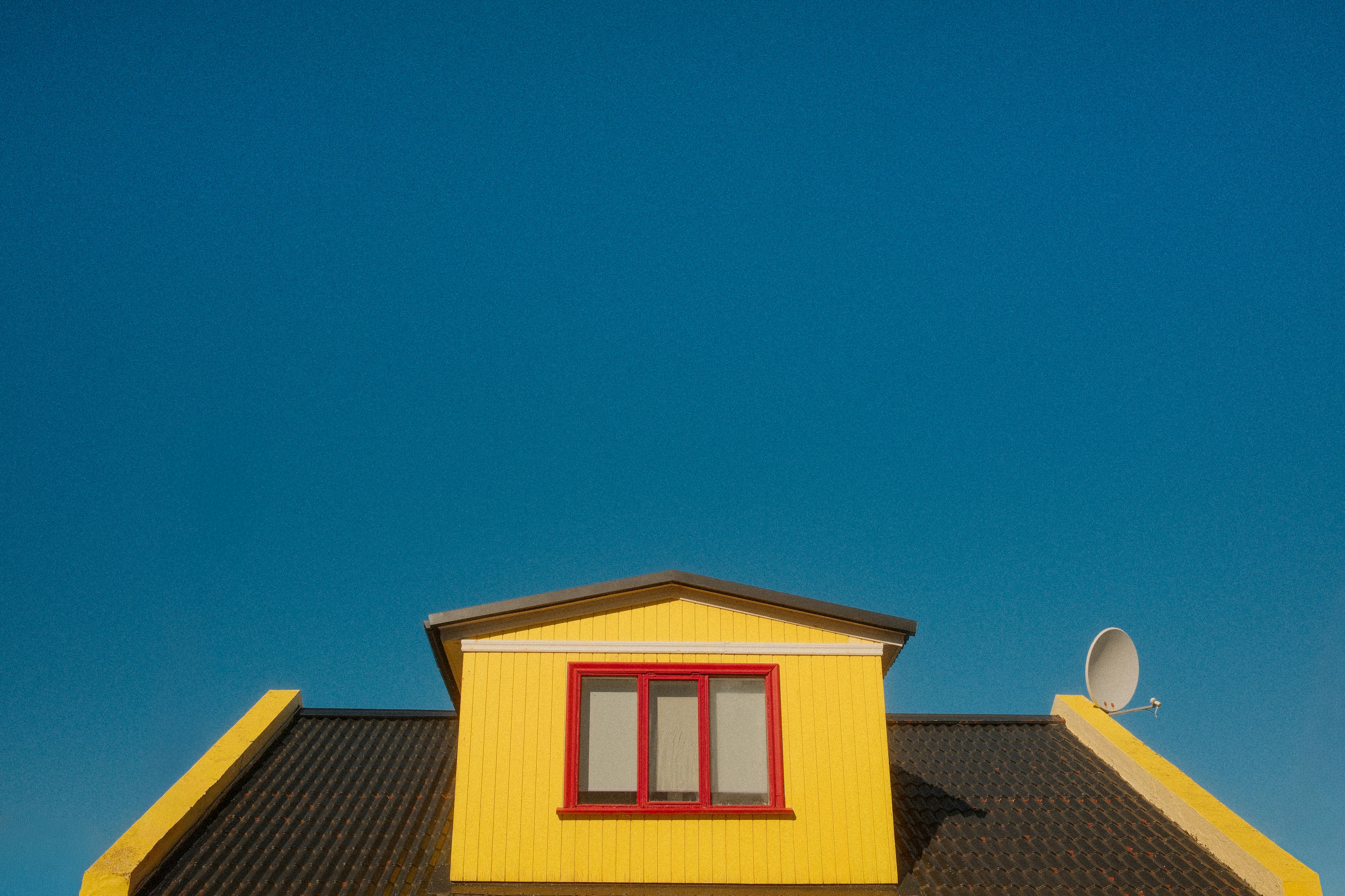 A yellow dormer with a red window frame on a dark roof against blue sky