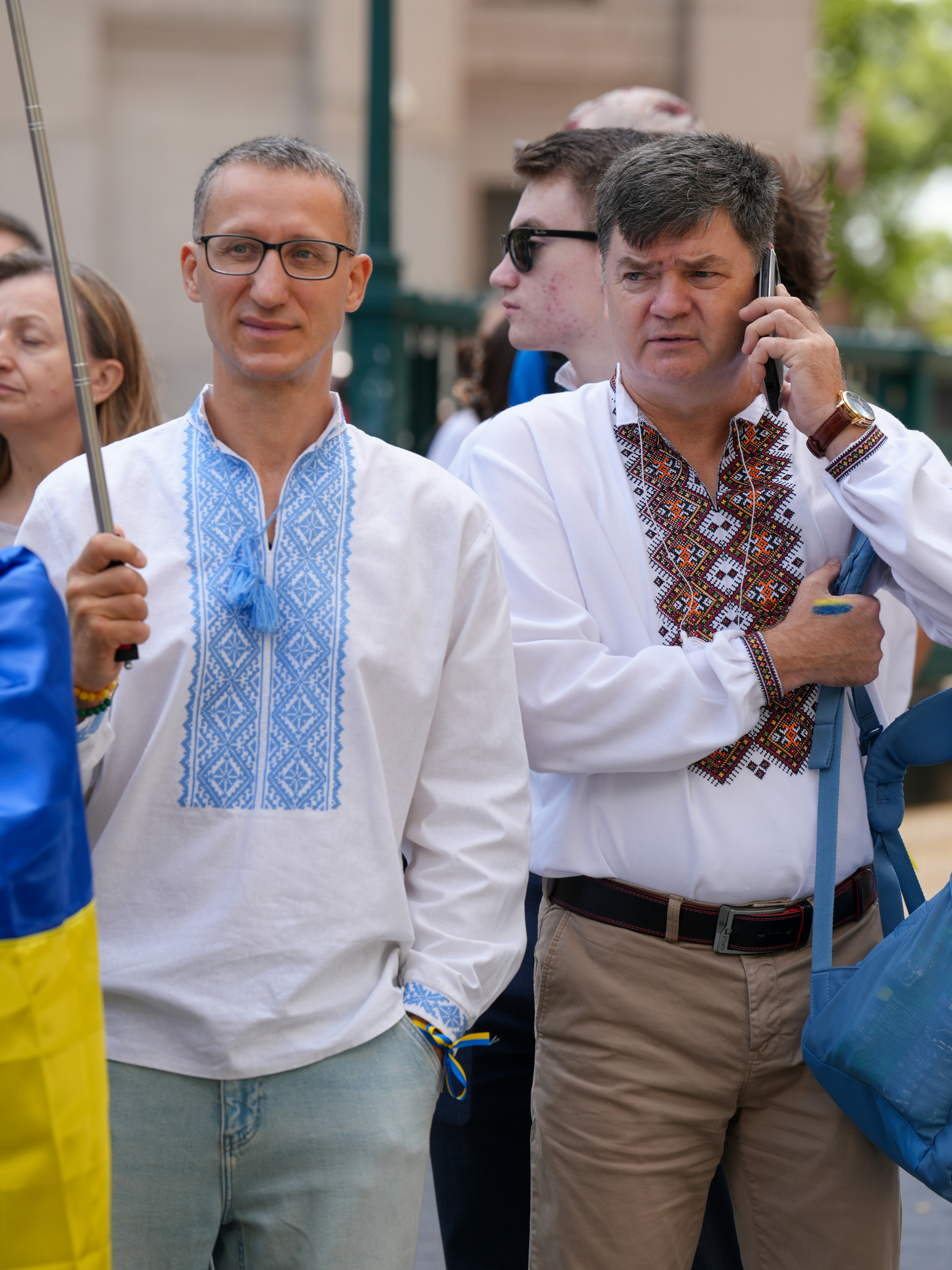 Two men wearing traditional embroidered shirts with a blue and yellow flag