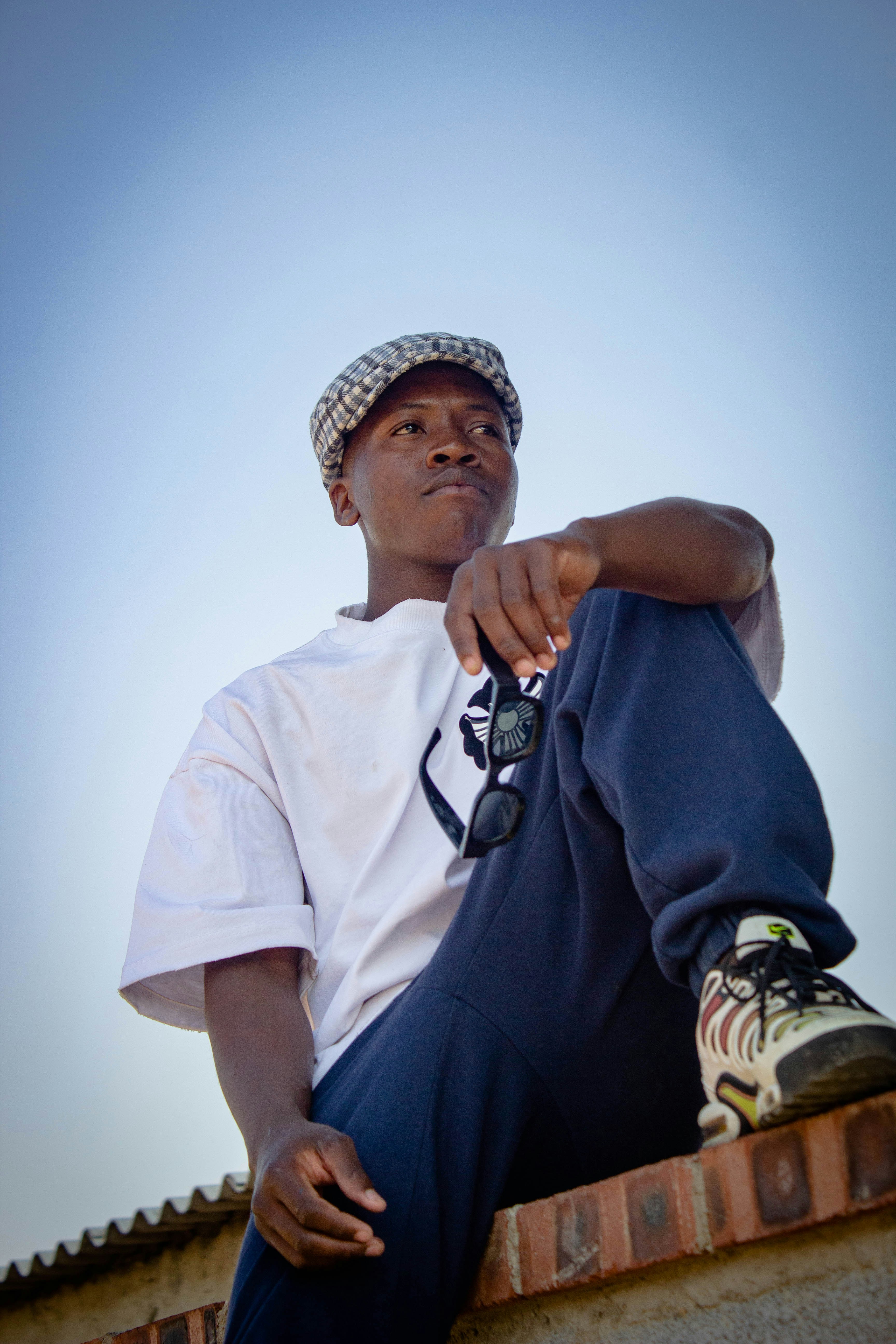 A young man in a plaid cap and white t-shirt holding sunglasses outdoors