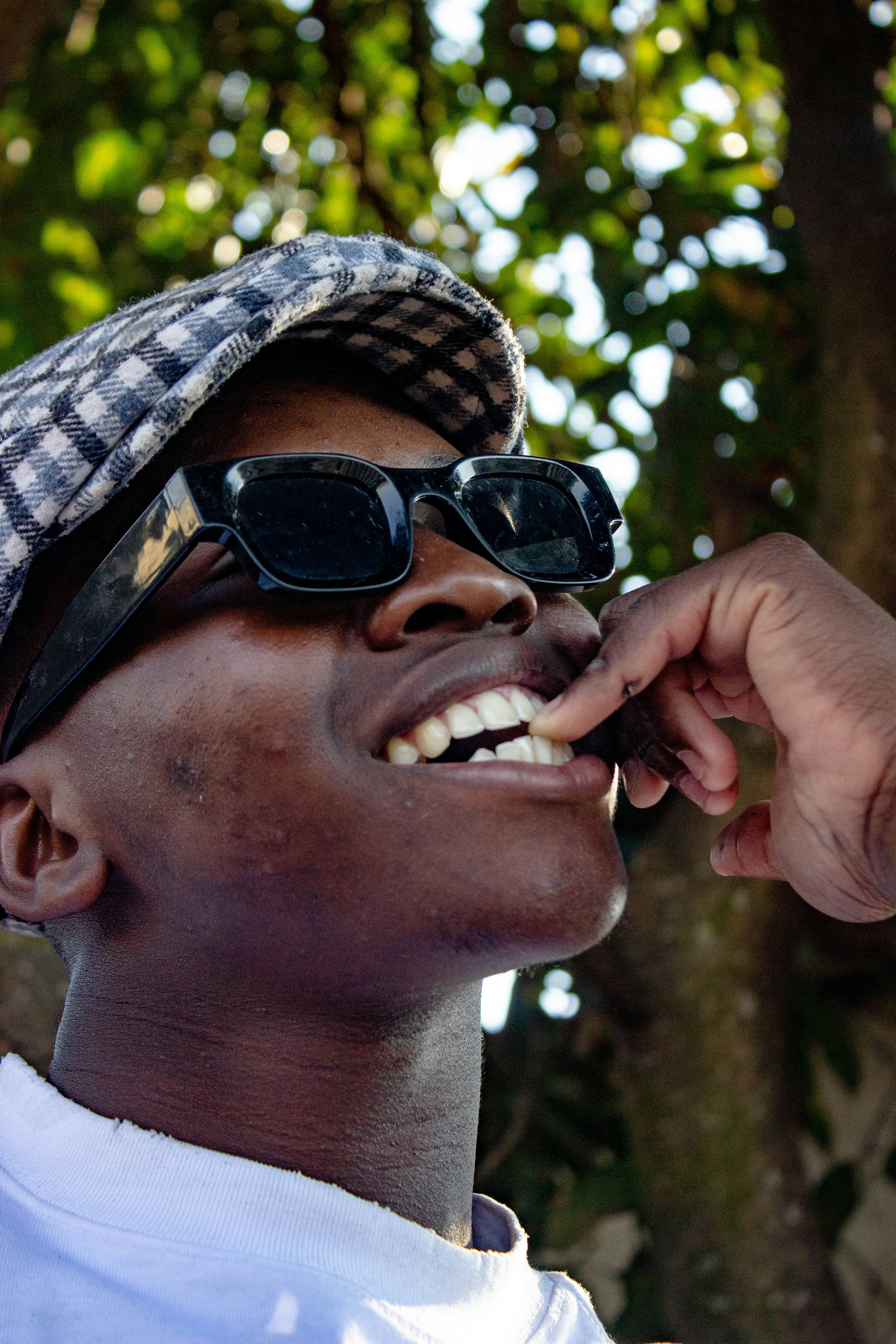 A person wearing a plaid flat cap and sunglasses touching their teeth